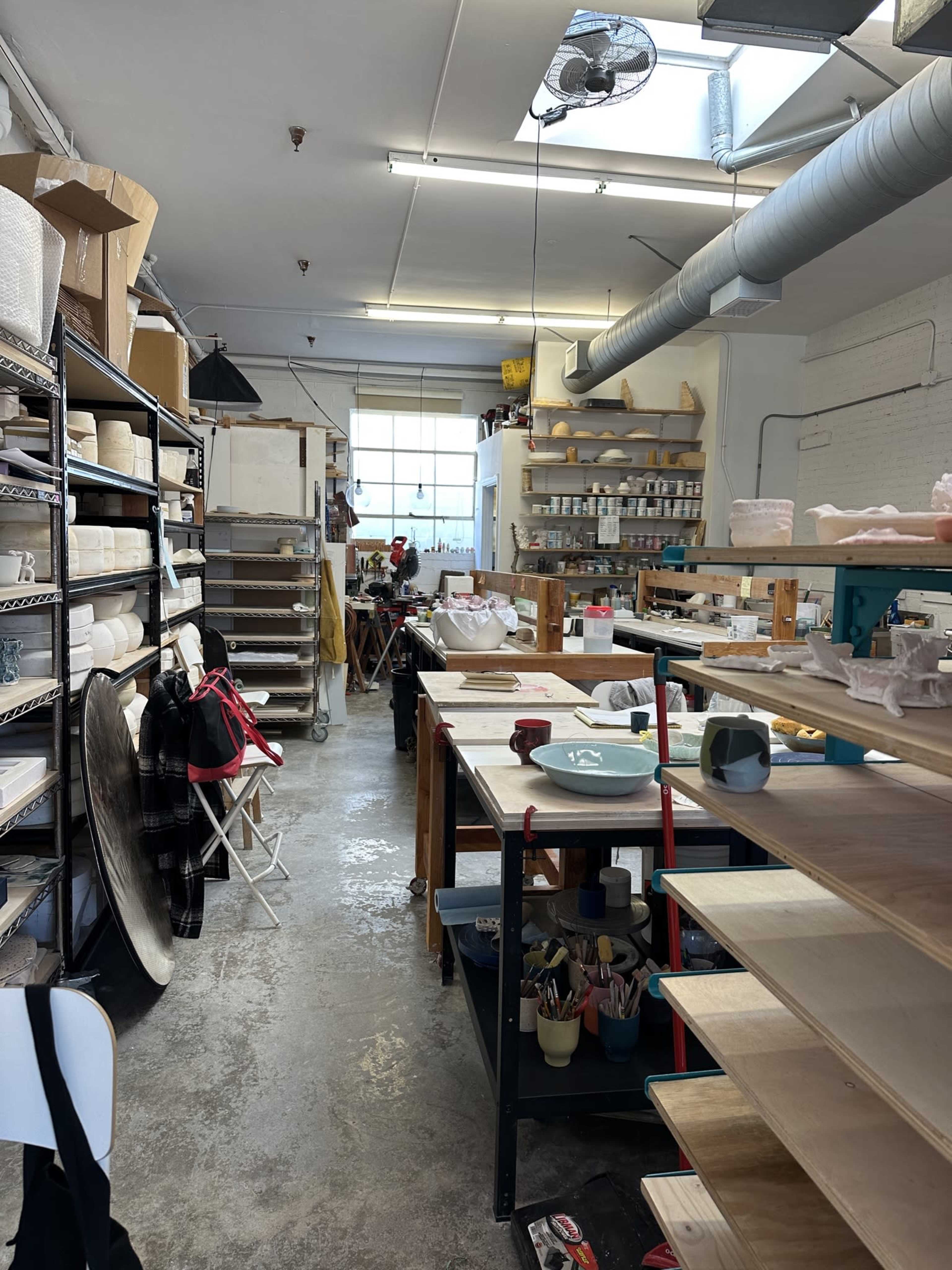 The image shows a ceramics studio with shelves filled with pottery supplies, work tables, and equipment arranged in a well-lit space.