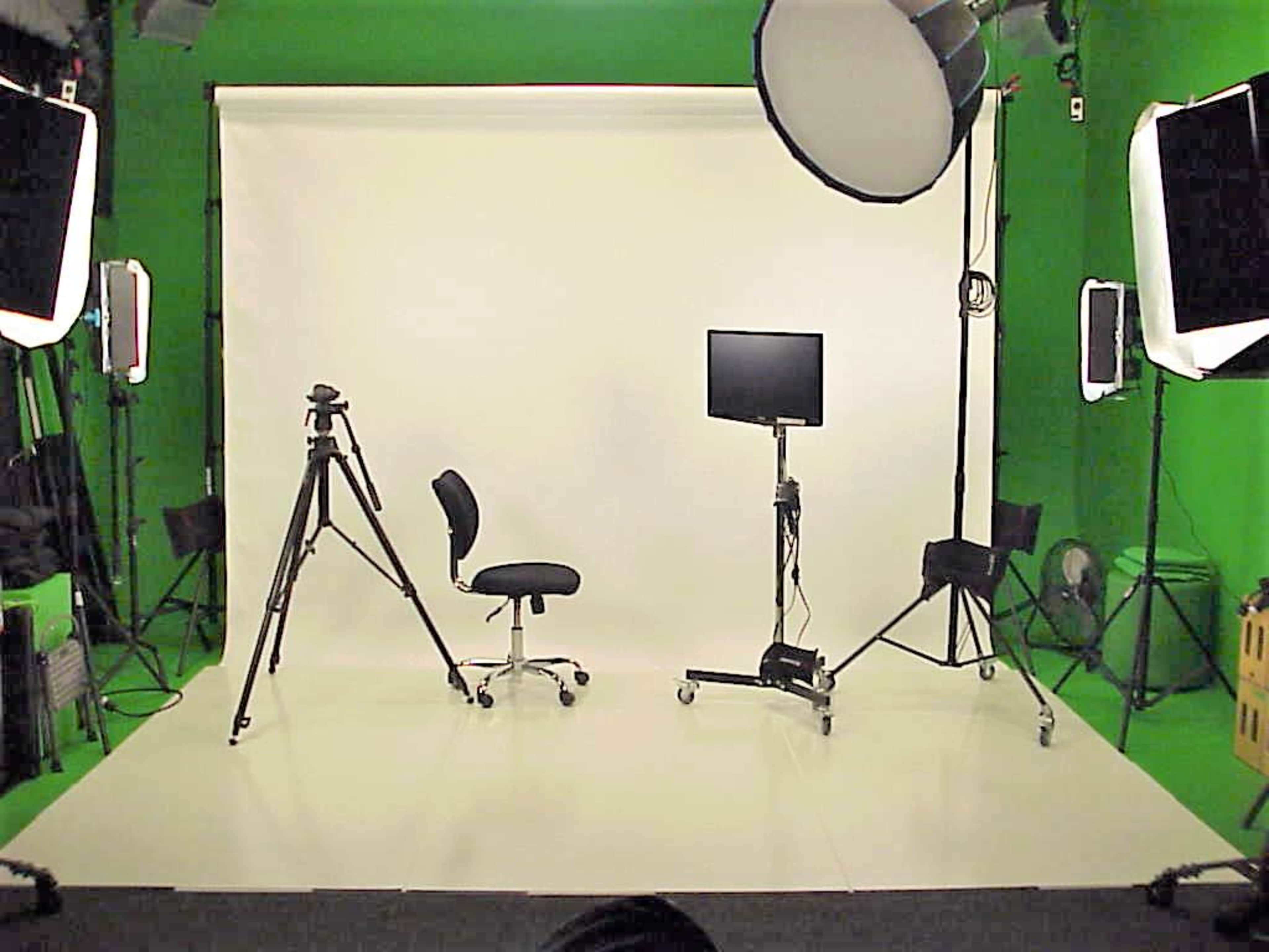 The image shows a photography studio setup with a white backdrop, a chair, a monitor on a wheeled stand, a tripod, and studio lights.