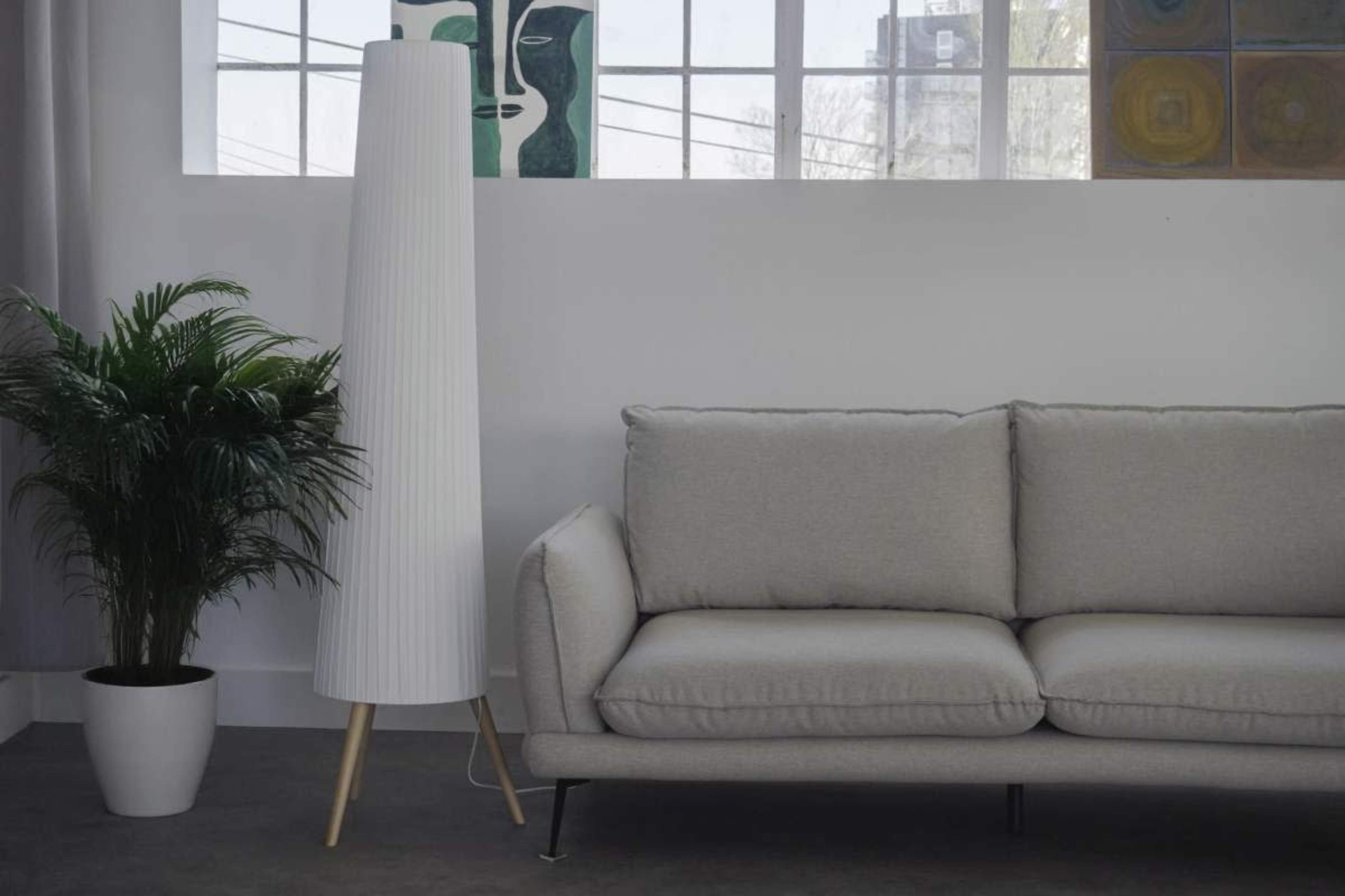 A modern living room features a light gray sofa, a tall white floor lamp, and a potted plant next to a window with an urban view.