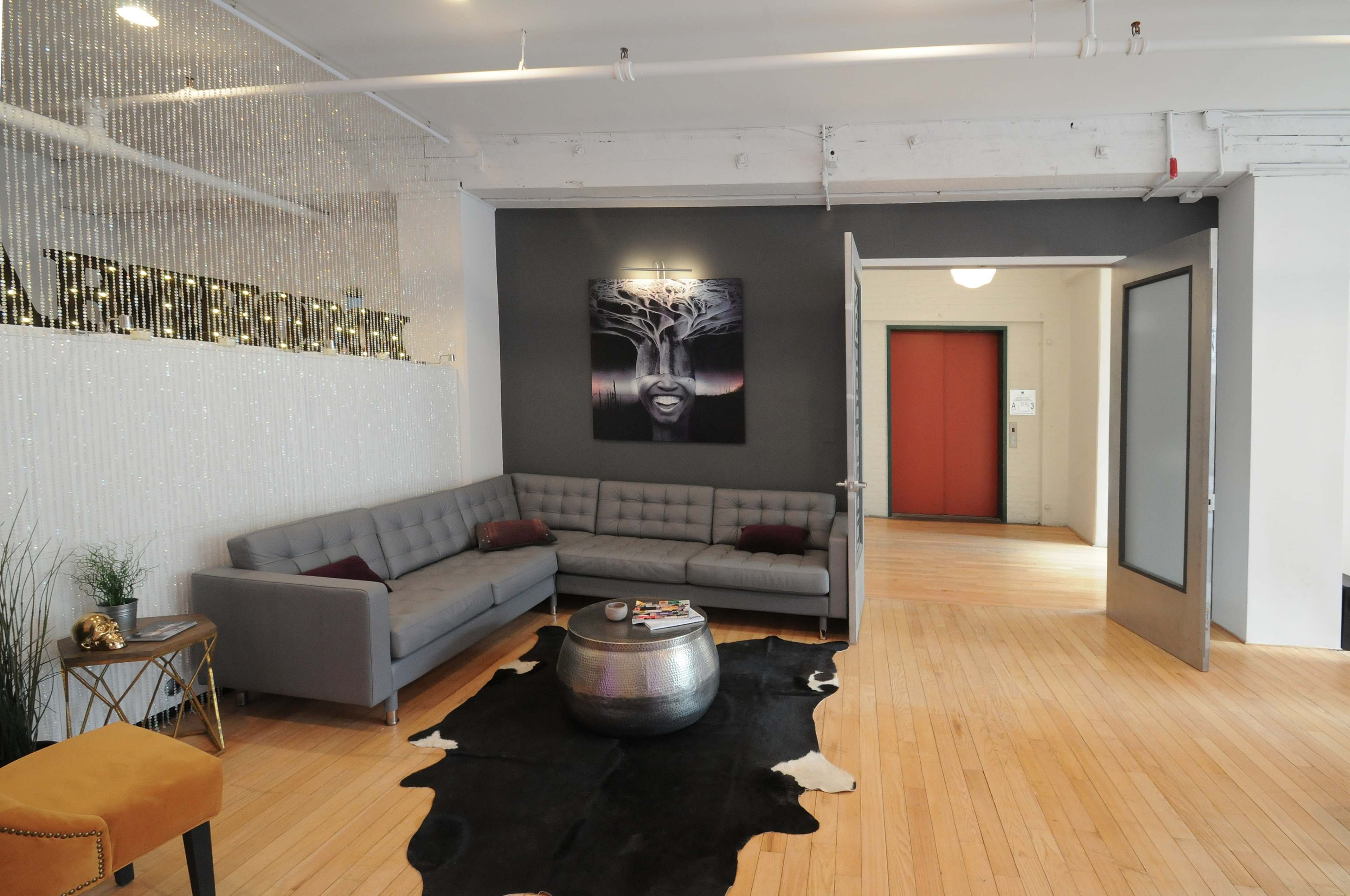 A modern lounge area with a gray sectional sofa, a round metallic coffee table, and a large artwork featuring a face on the wall, adjacent to an entrance with red elevator doors.