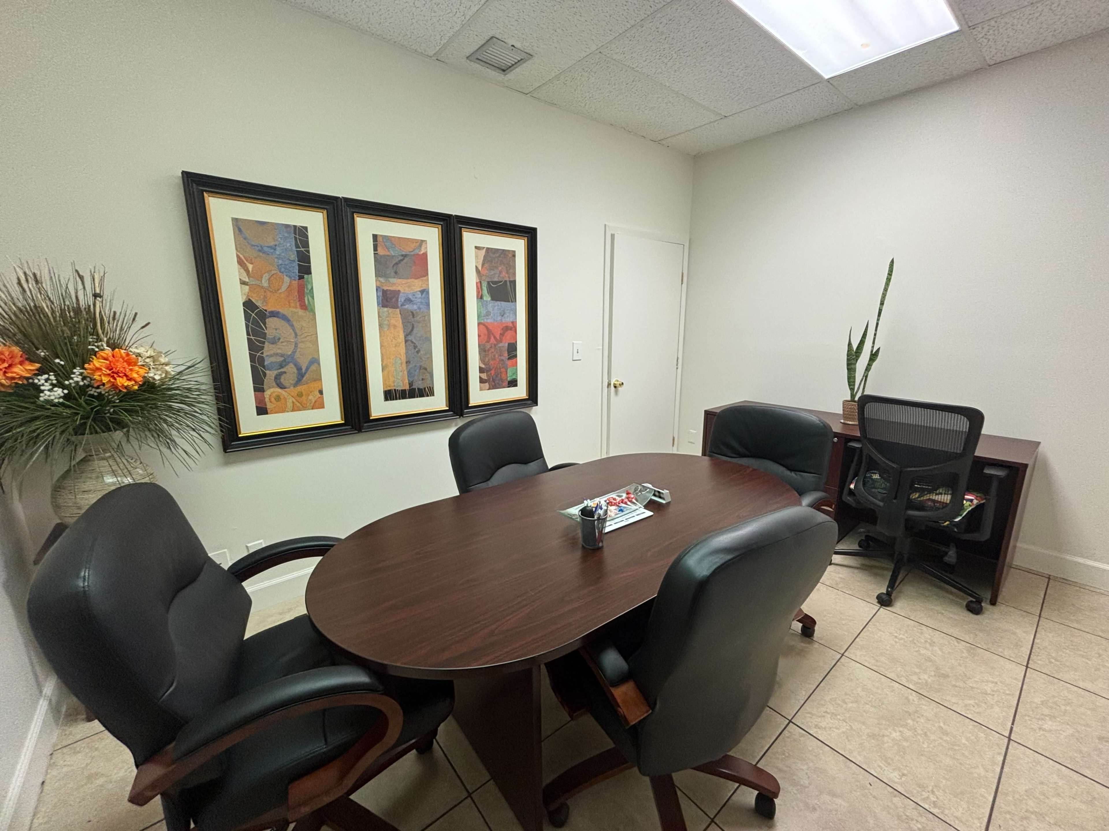 The image shows a small meeting room featuring a round wooden table surrounded by four black chairs, with two framed artworks on the wall and a small desk with a chair in the corner.