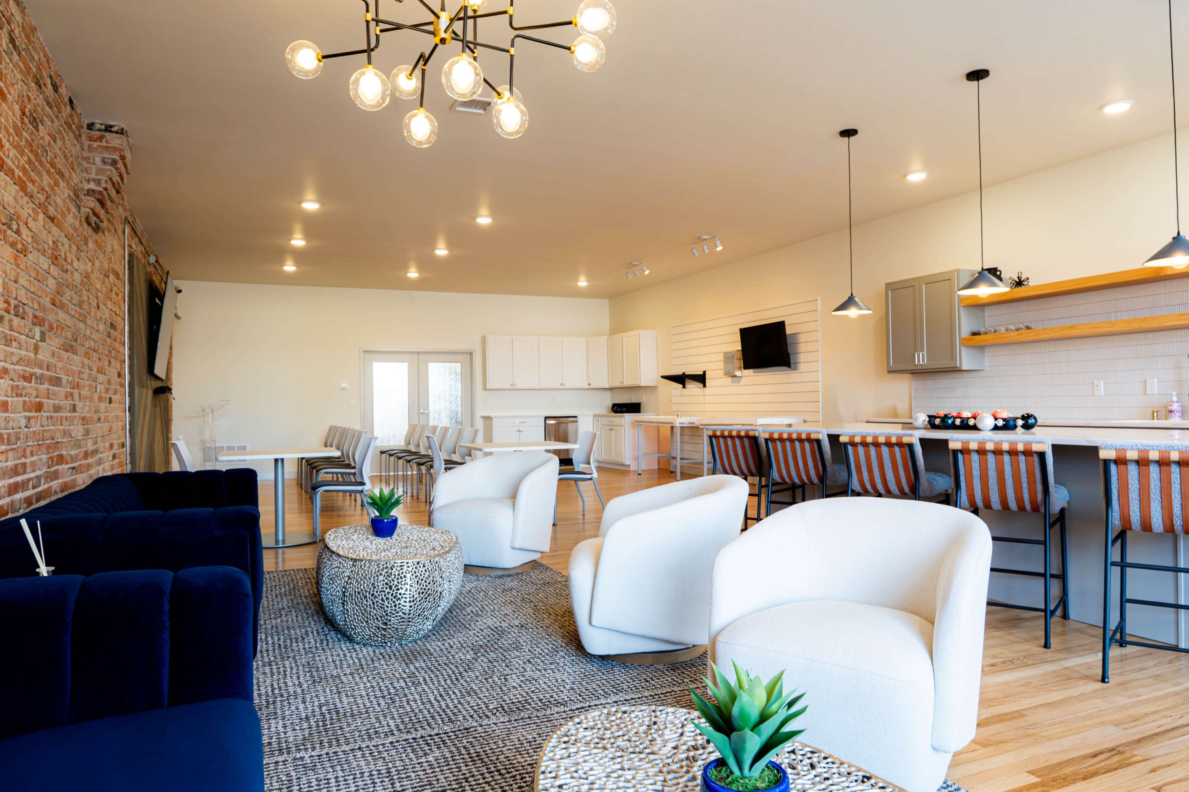 The image shows a modern communal living space with a mix of seating, a kitchen area, and decorative plants.