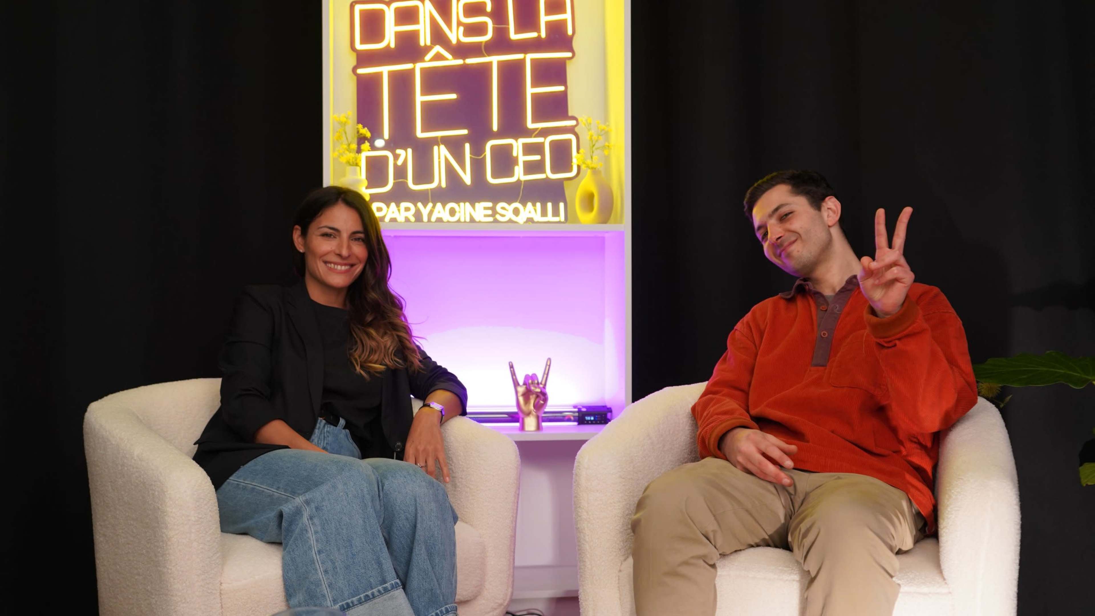 Two people are seated in modern chairs in front of a neon sign that reads "DANS LA TÊTE D'UN CEO" in a well-lit studio setting.