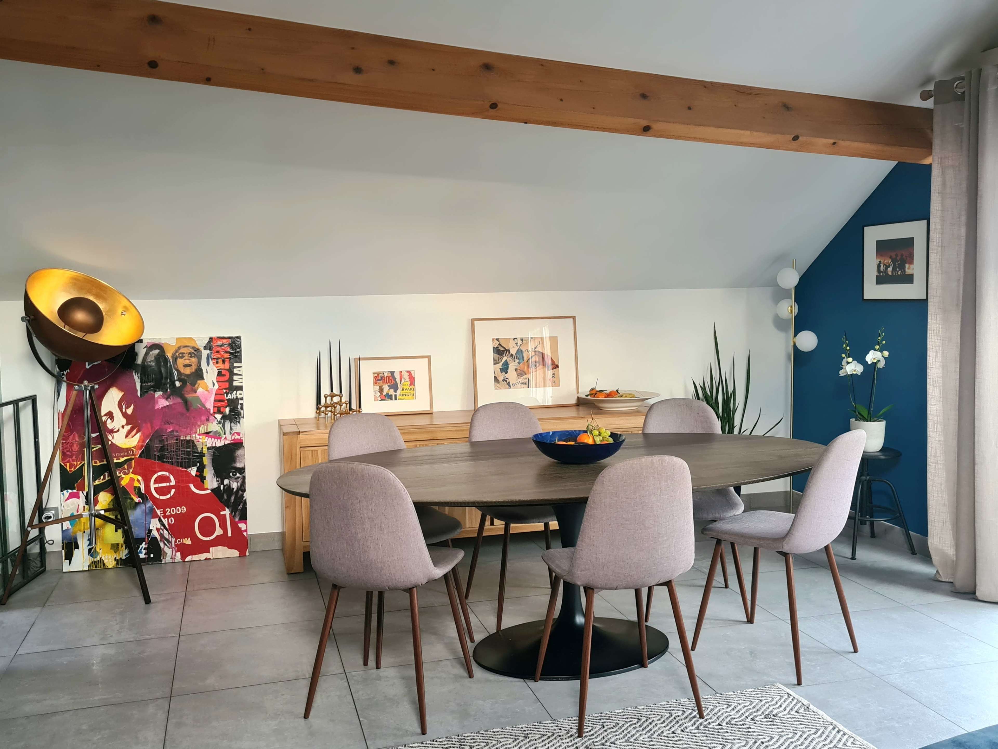 A modern dining area features a round table surrounded by six gray chairs, accompanied by a wooden sideboard and colorful artwork on the walls.