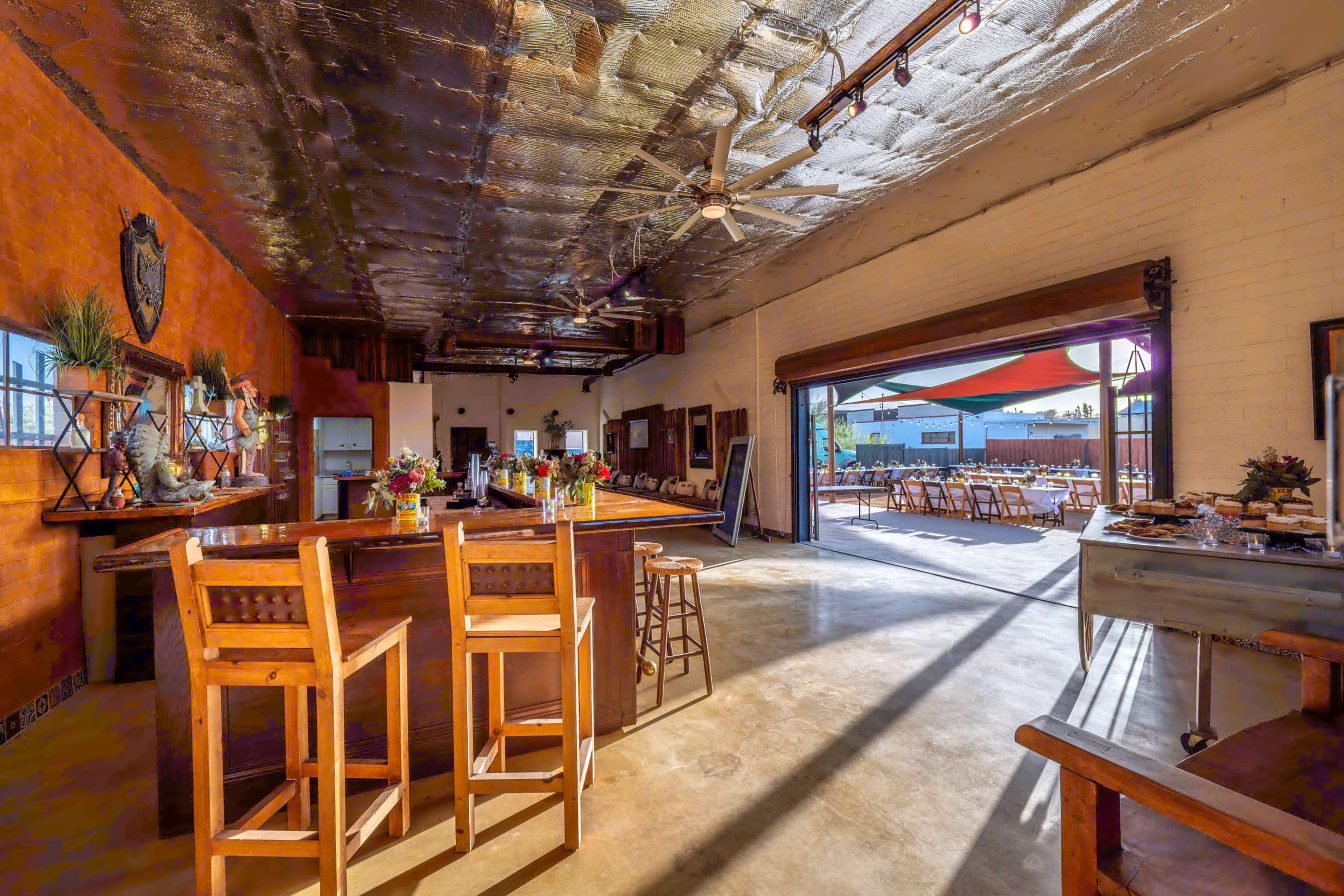 The image shows a spacious interior of a bar with wooden furniture, large windows, and an adjacent outdoor seating area.