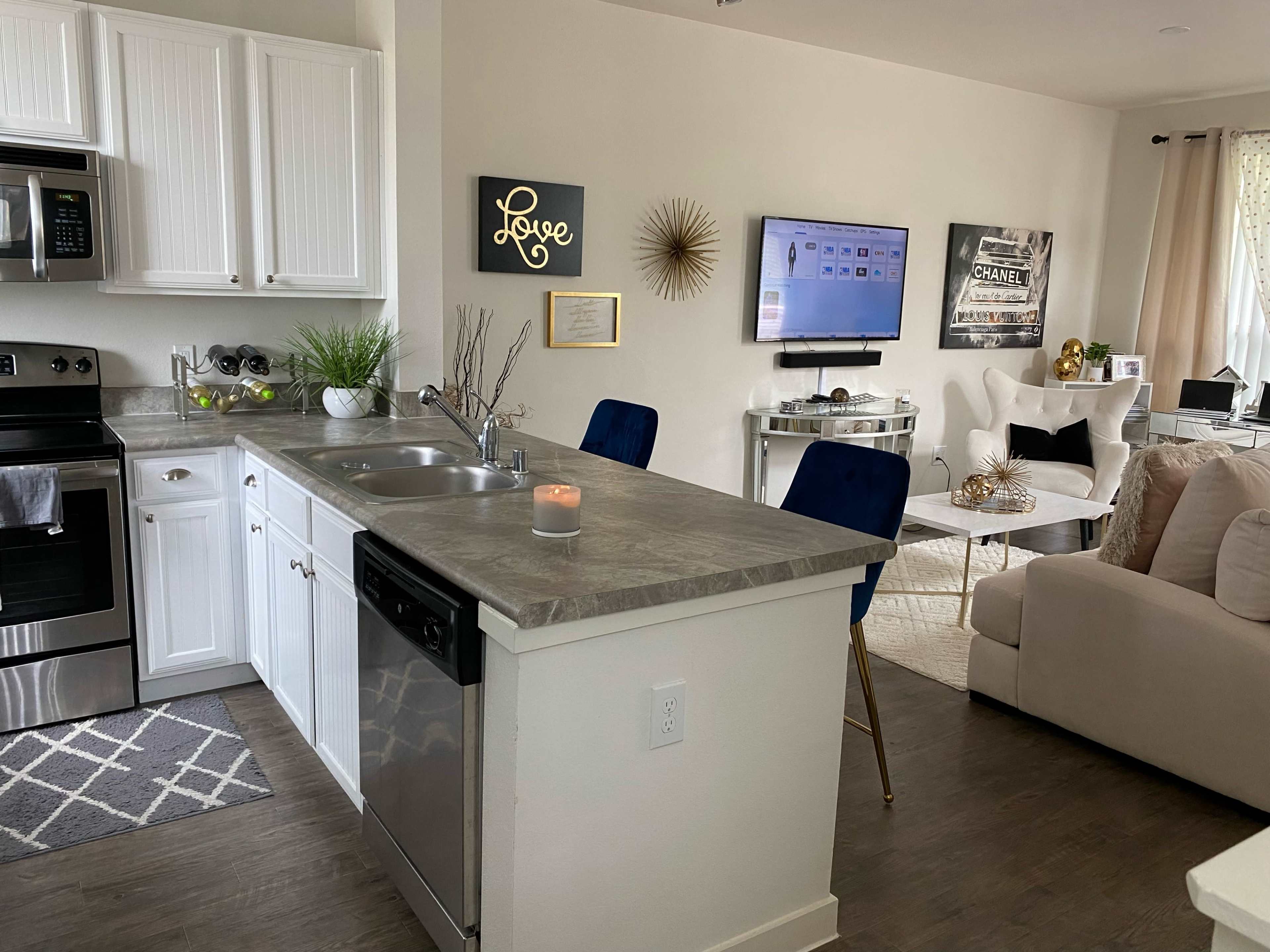 A modern kitchen with white cabinets, a gray countertop, and a double sink, featuring a small dining area with blue chairs and a living space with a television, decorative items, and a beige sofa.