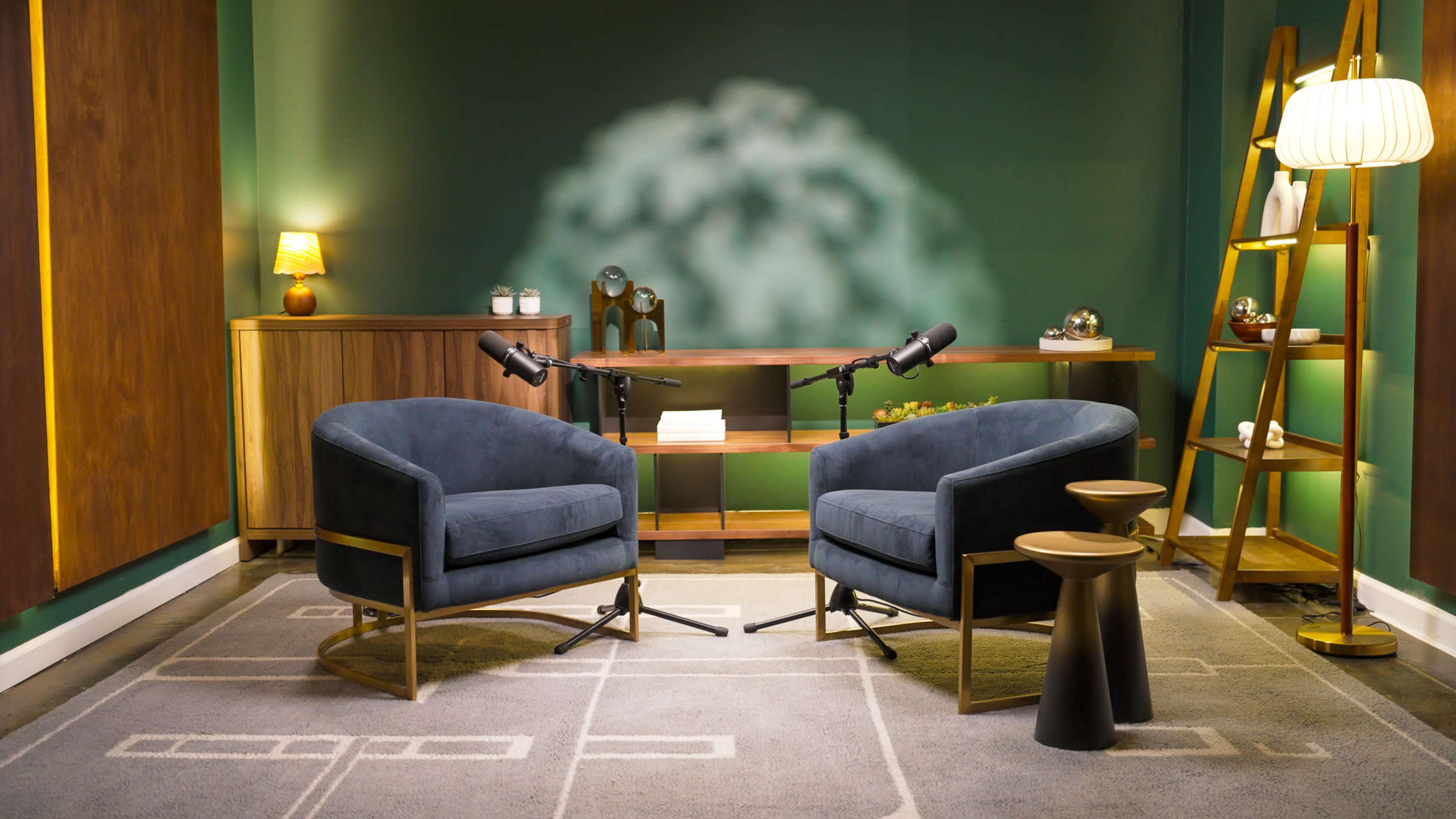 The image features a modern interview setup with two velvet chairs, microphones positioned on stands, a wooden shelf with decorative items, and a green wall backdrop.