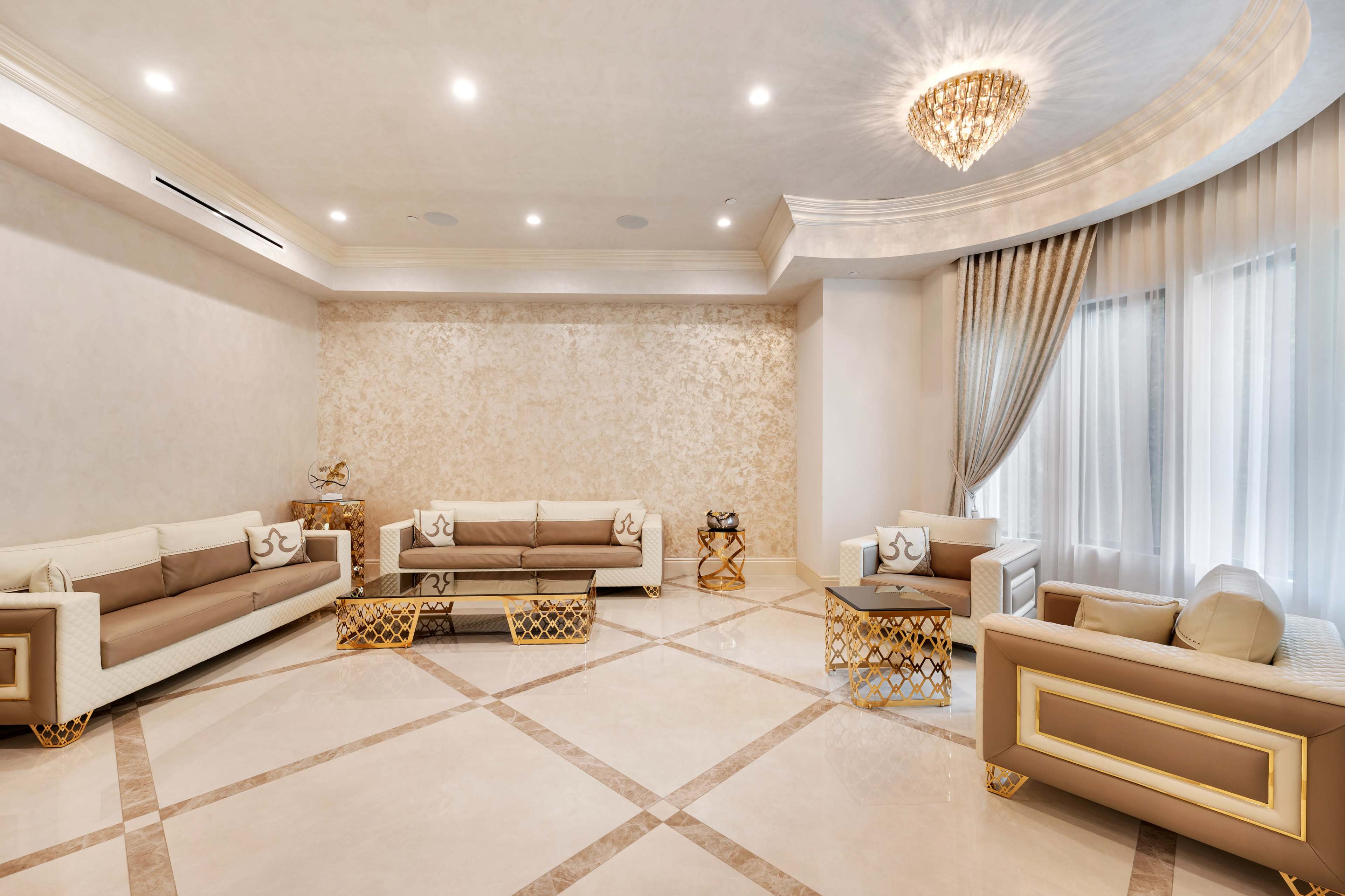 The image shows a spacious and elegantly designed living room with cream-colored sofas, glass-topped tables, and a decorative chandelier.