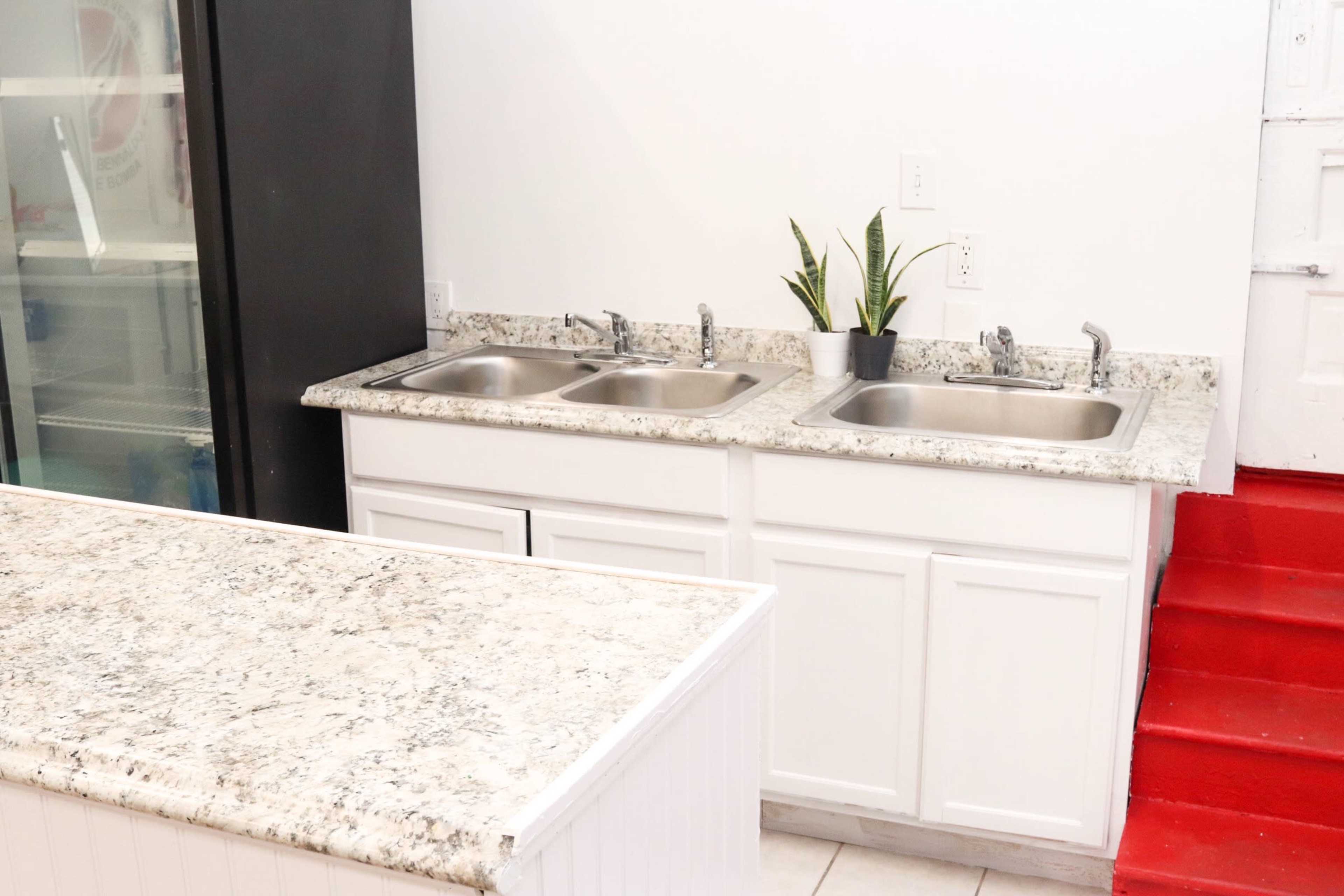 A kitchen area featuring a white countertop with two metal sinks and a small potted plant, alongside a black refrigerator and red stairs.