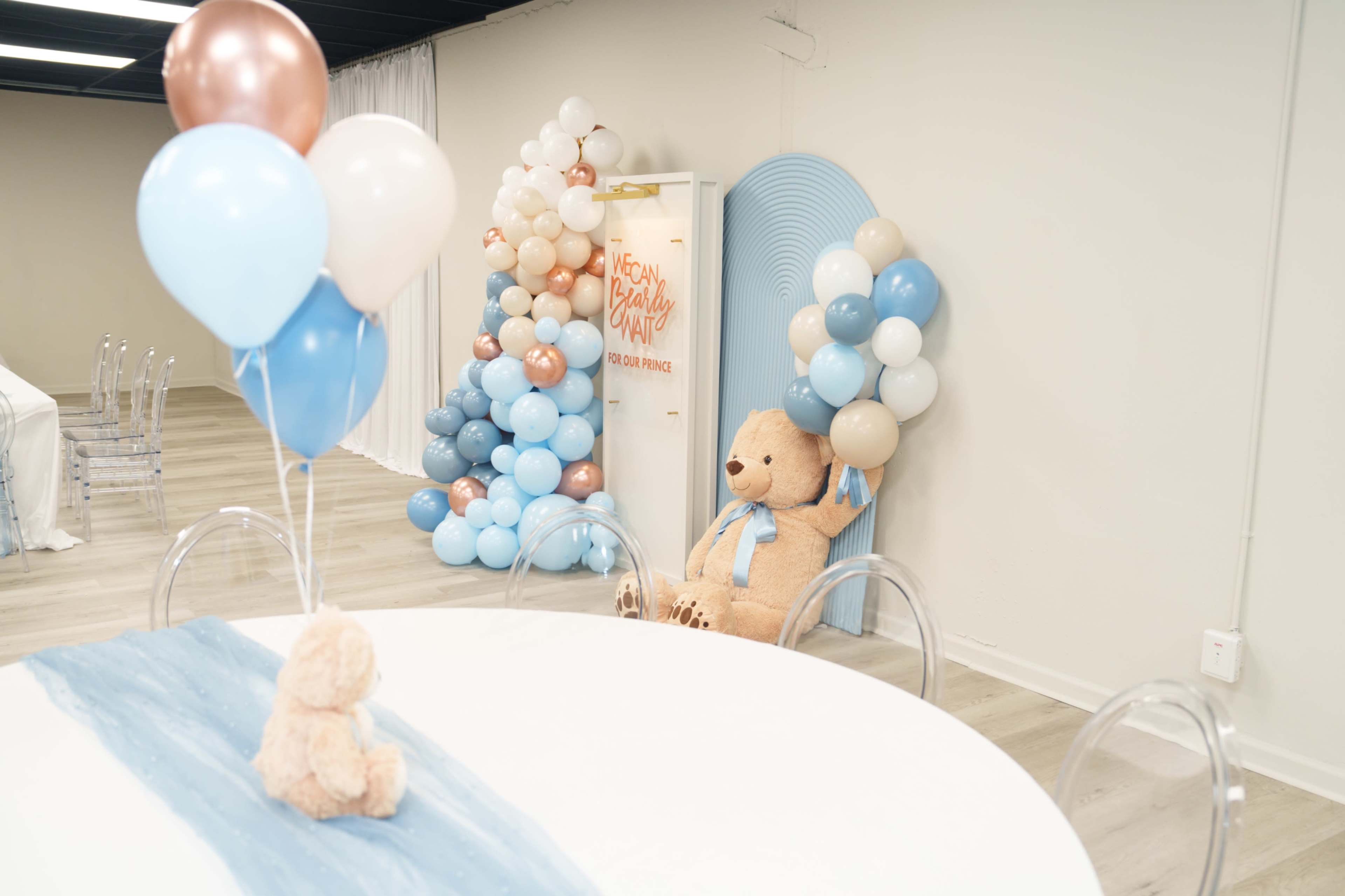 The image shows a decorated room featuring a large teddy bear, a table with a light blue runner, and colorful balloons arranged on one wall.