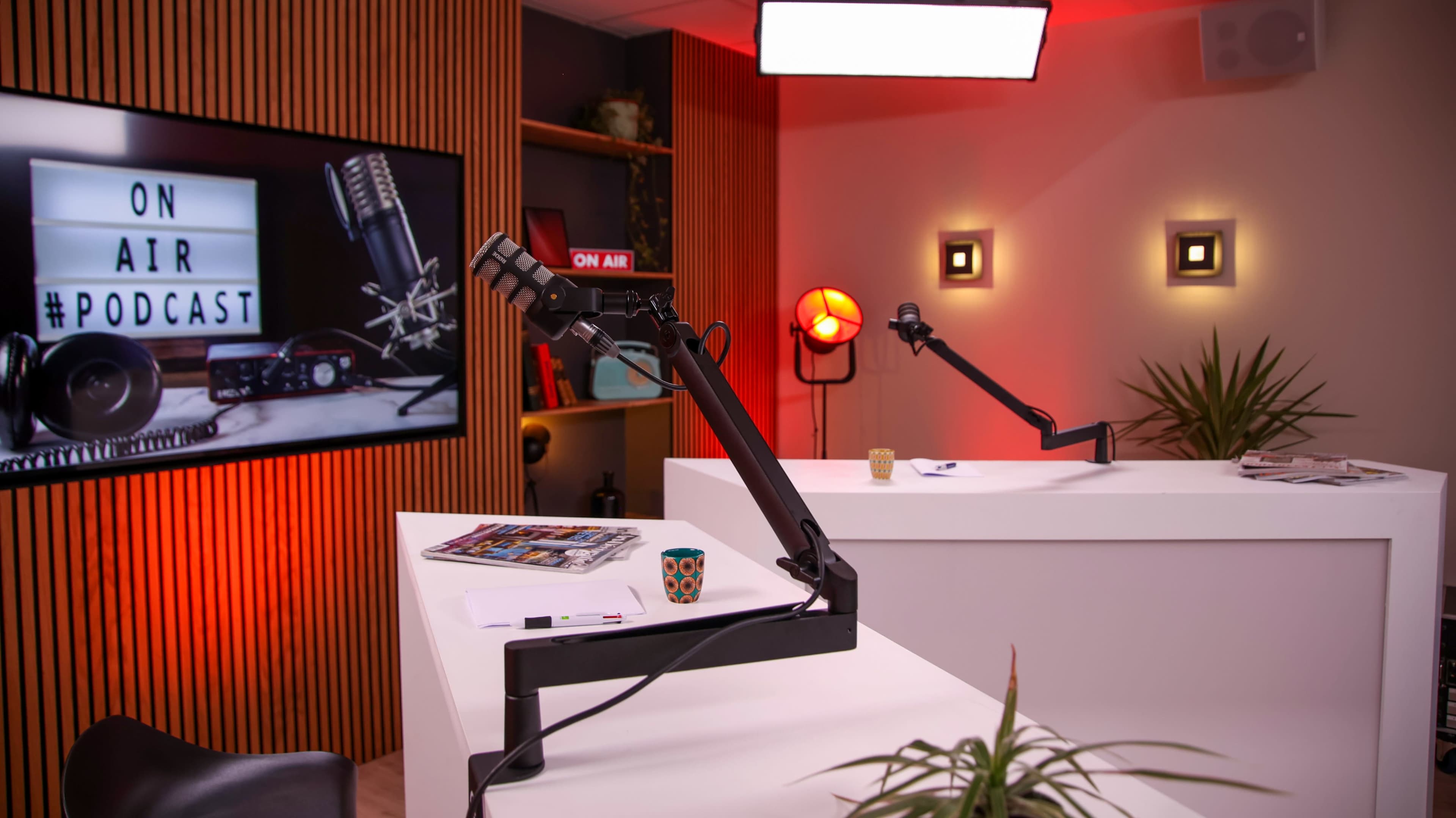 The image shows a modern podcast studio with a microphone on a boom arm, a large screen displaying "ON AIR #PODCAST," and two workstations with plants and a coffee cup.