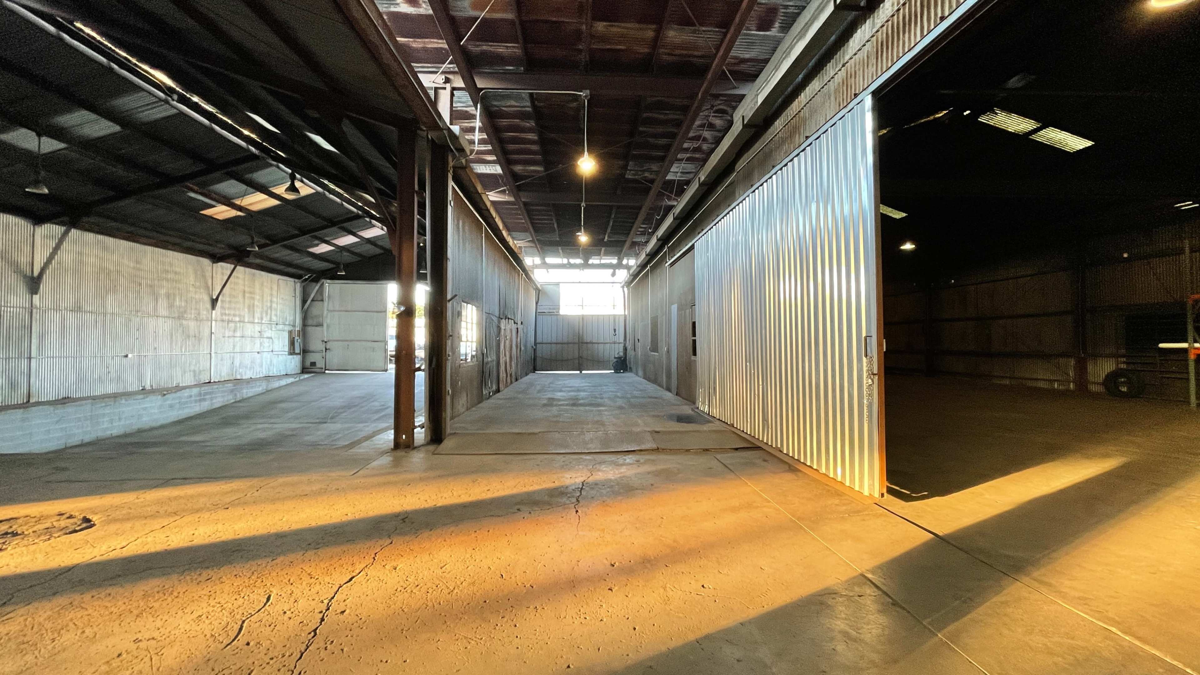 A large, industrial warehouse interior with open doors allowing light to stream in, illuminating the concrete floor and metal structures.