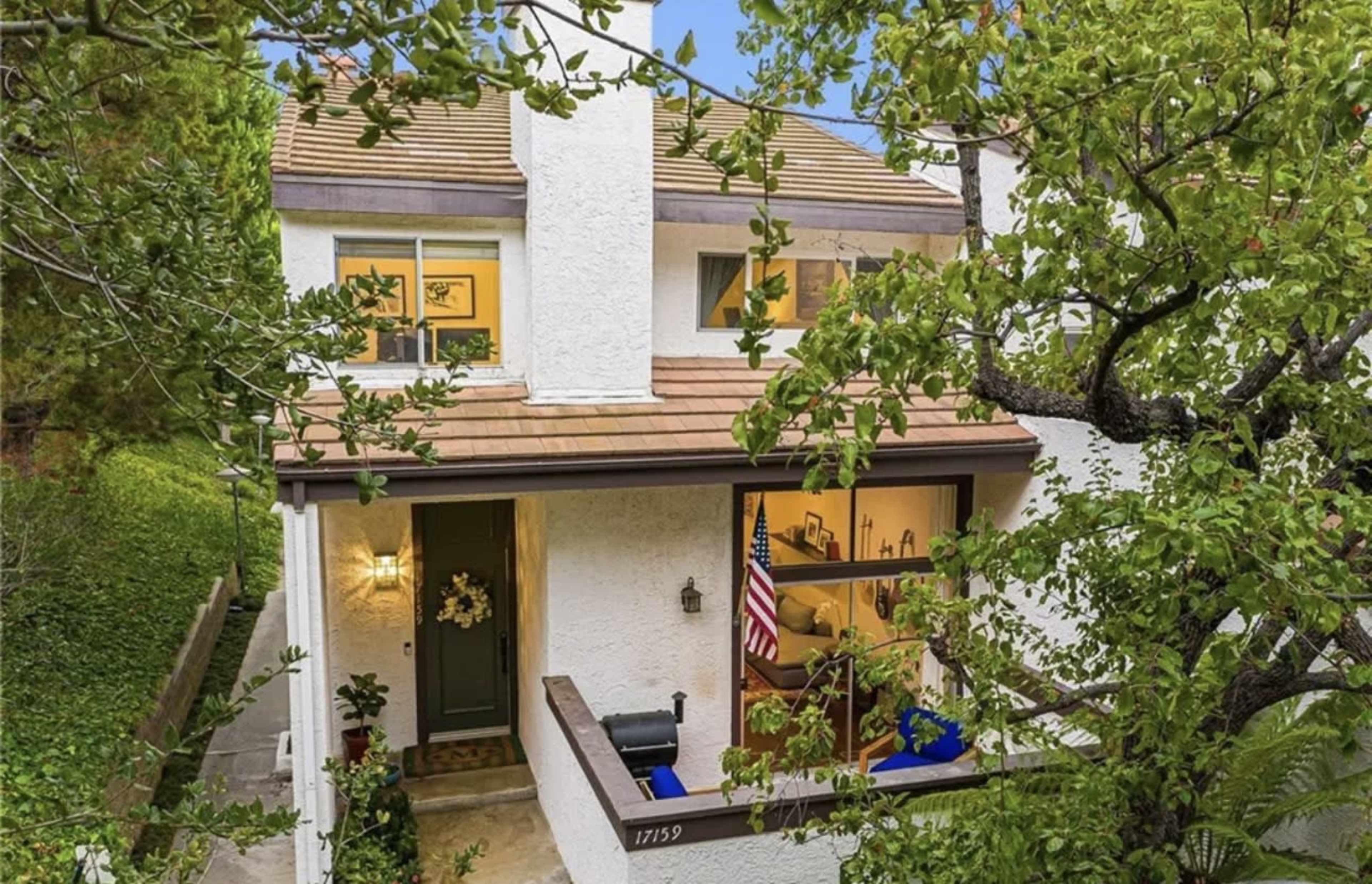 A two-story white house with a brown roof and an American flag visible through a window.