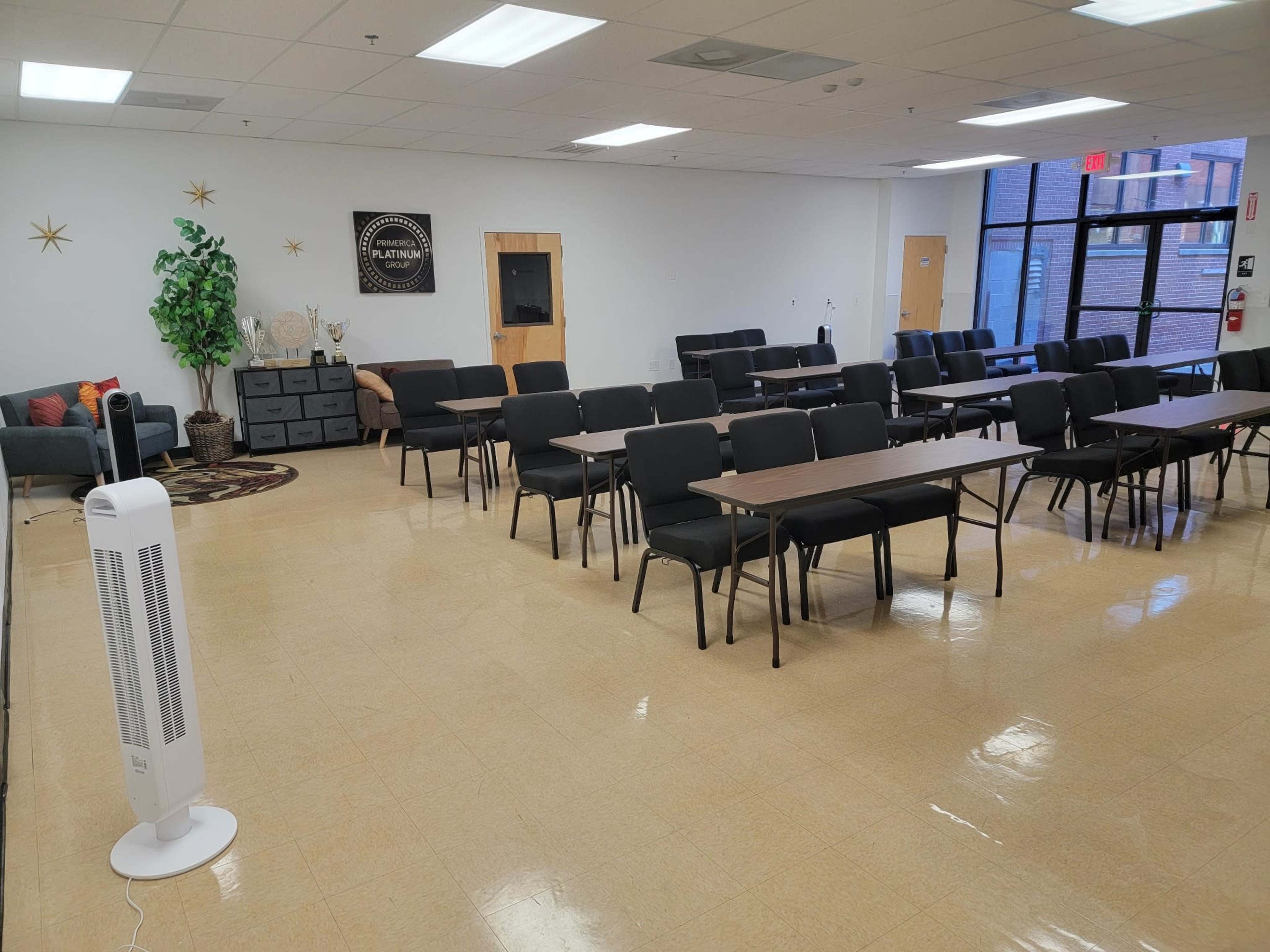 The image shows a spacious room set up with multiple rows of tables and chairs, a seating area to the left, and large windows providing natural light.