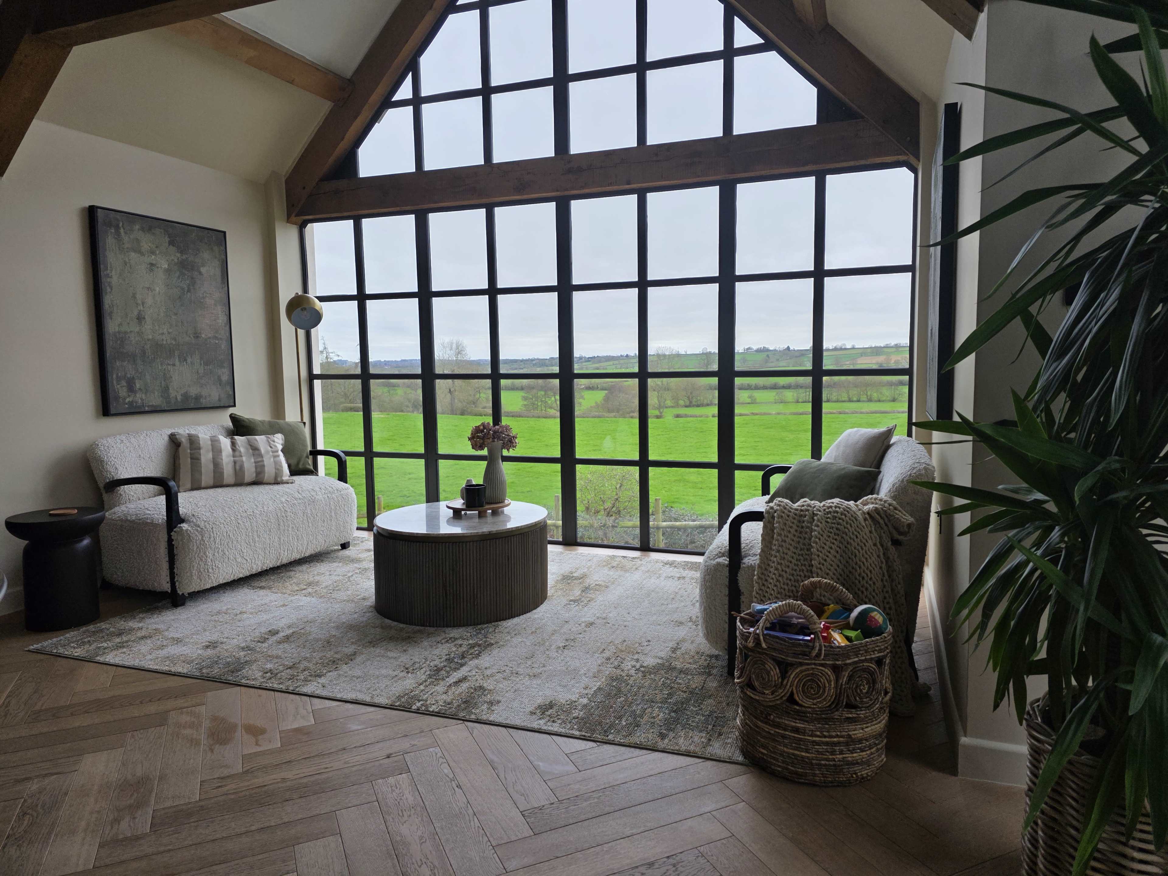 The image shows a cozy living space with two armchairs, a round coffee table, a large window offering a view of a green landscape, and a decorative plant in the corner.