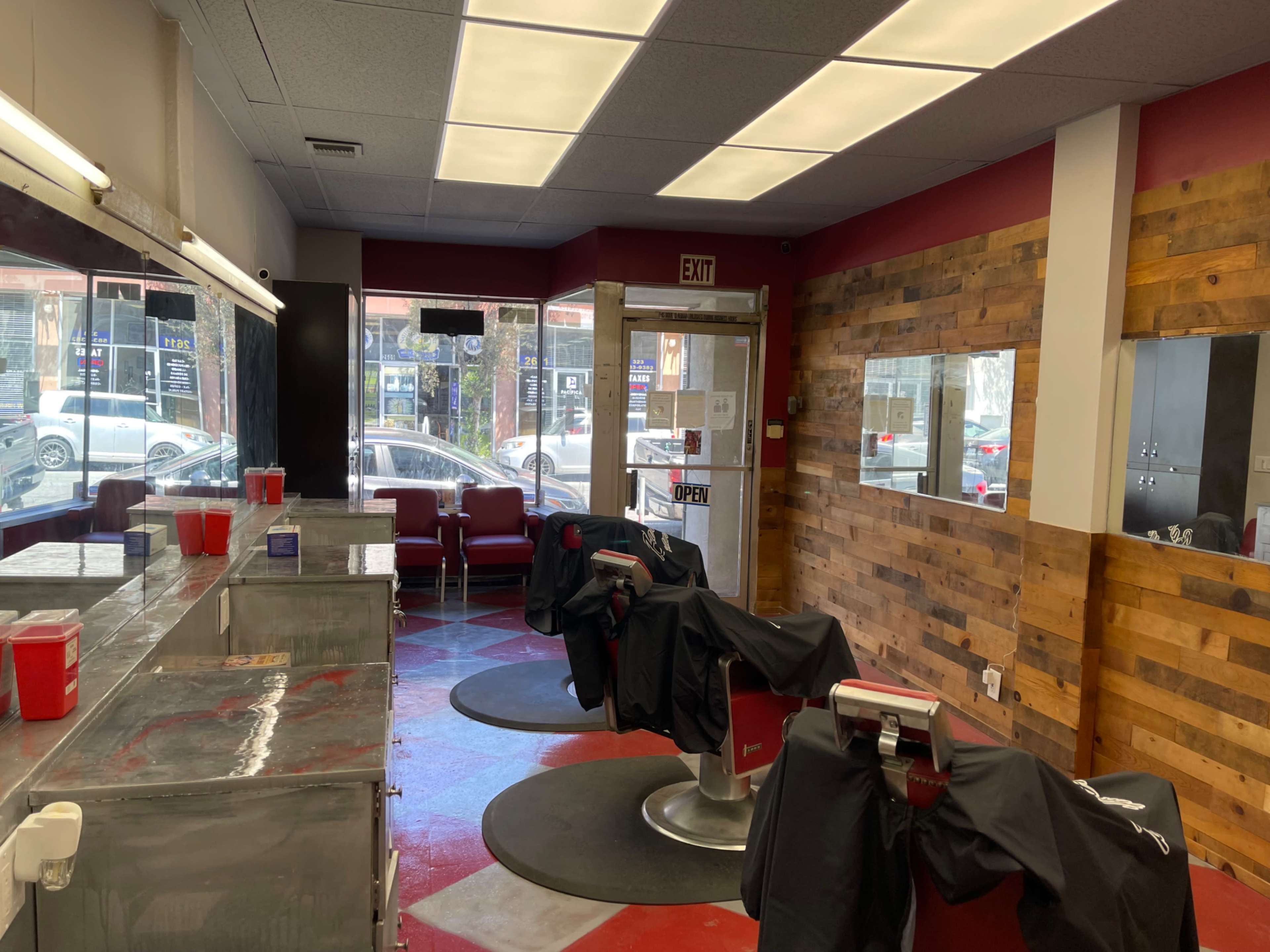The interior of a barbershop features three salon chairs with black capes, mirrored walls, and wooden accents, while an exit door leads to the street outside.