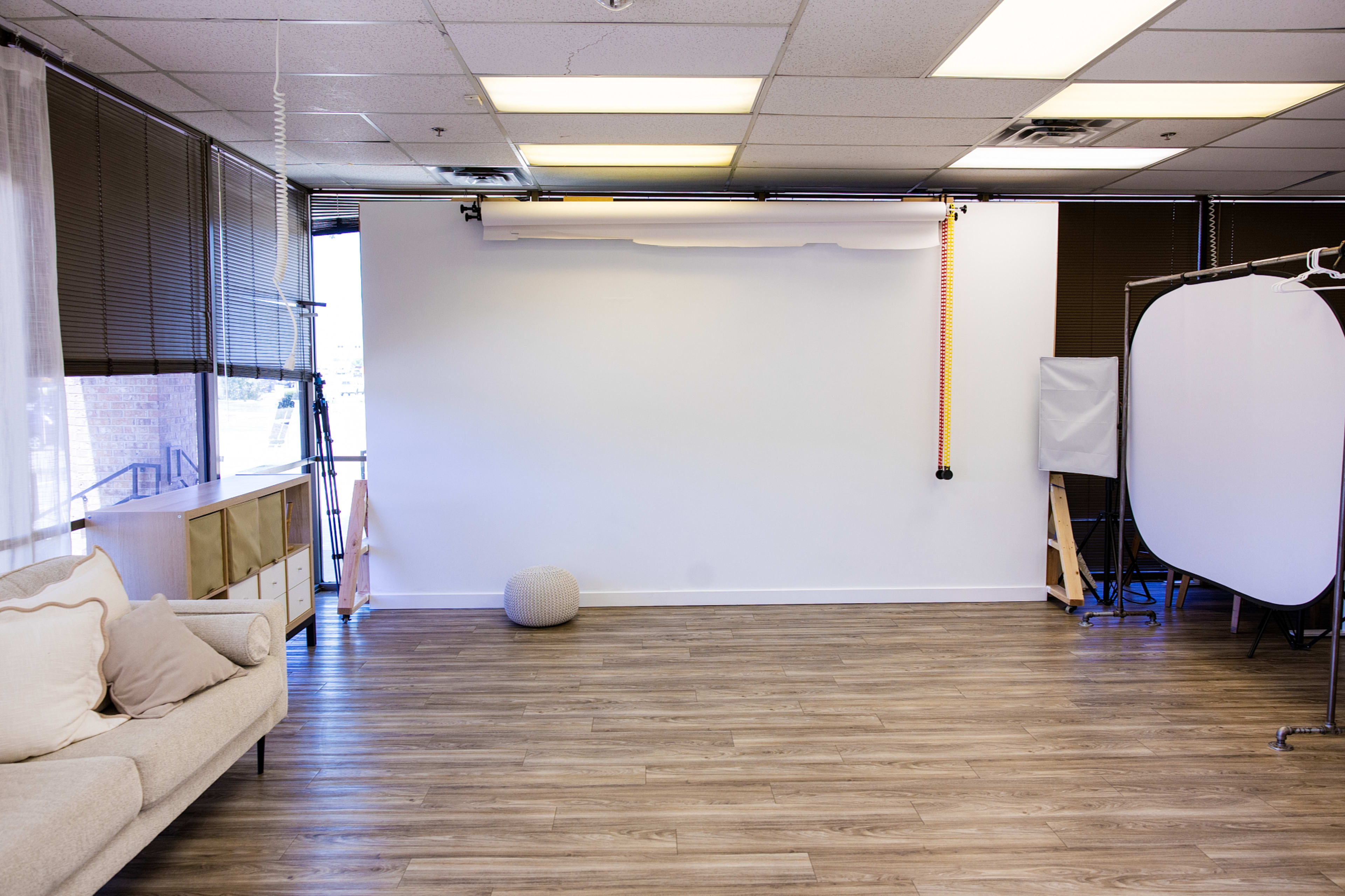 The image shows a minimalist studio space with a light-colored sofa, a large white backdrop, and various photography equipment arranged around the room.