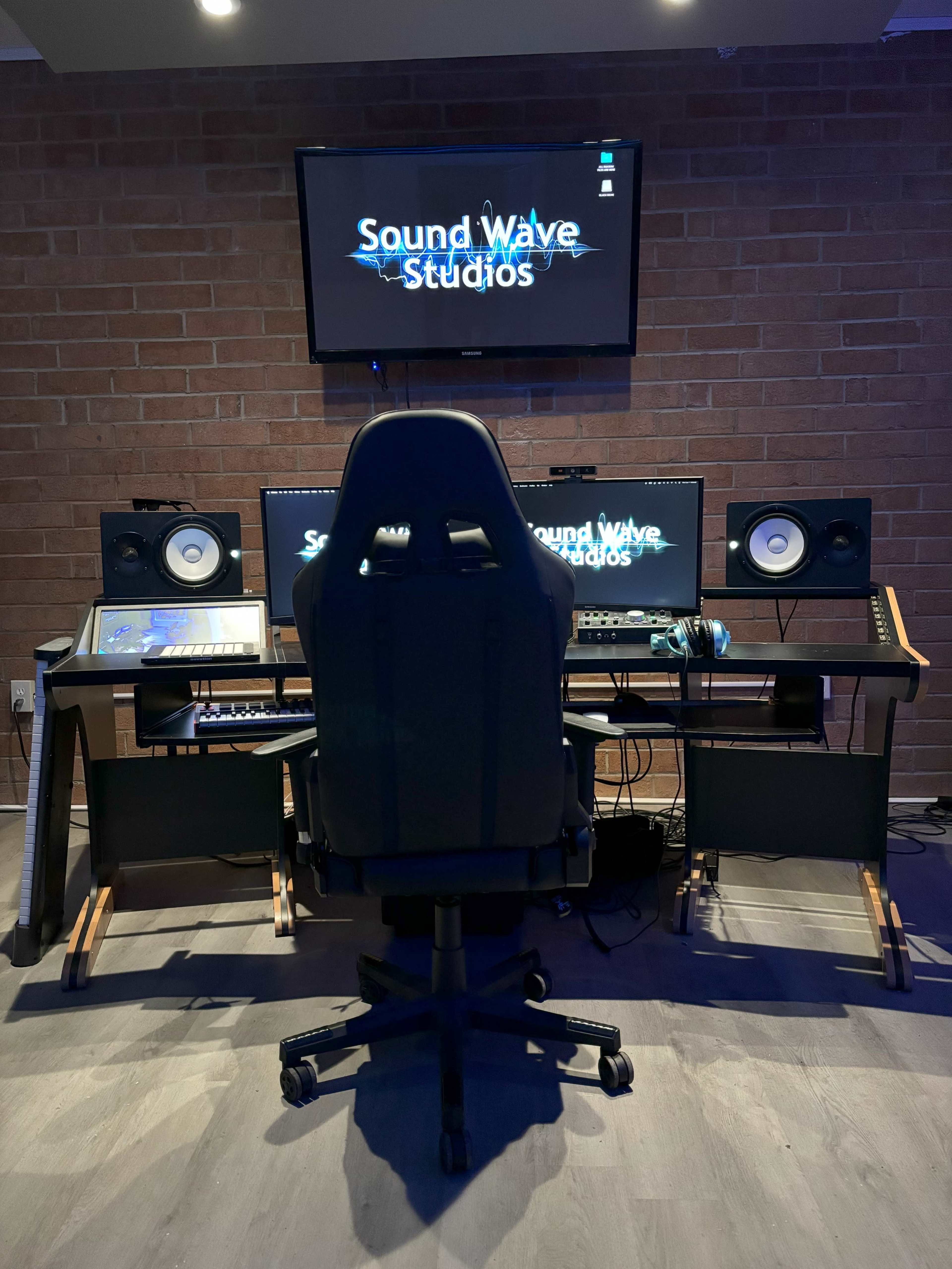 The image shows a music production studio setup with a desk, computer monitors, audio speakers, and a black ergonomic chair facing the screens, all against a brick wall.