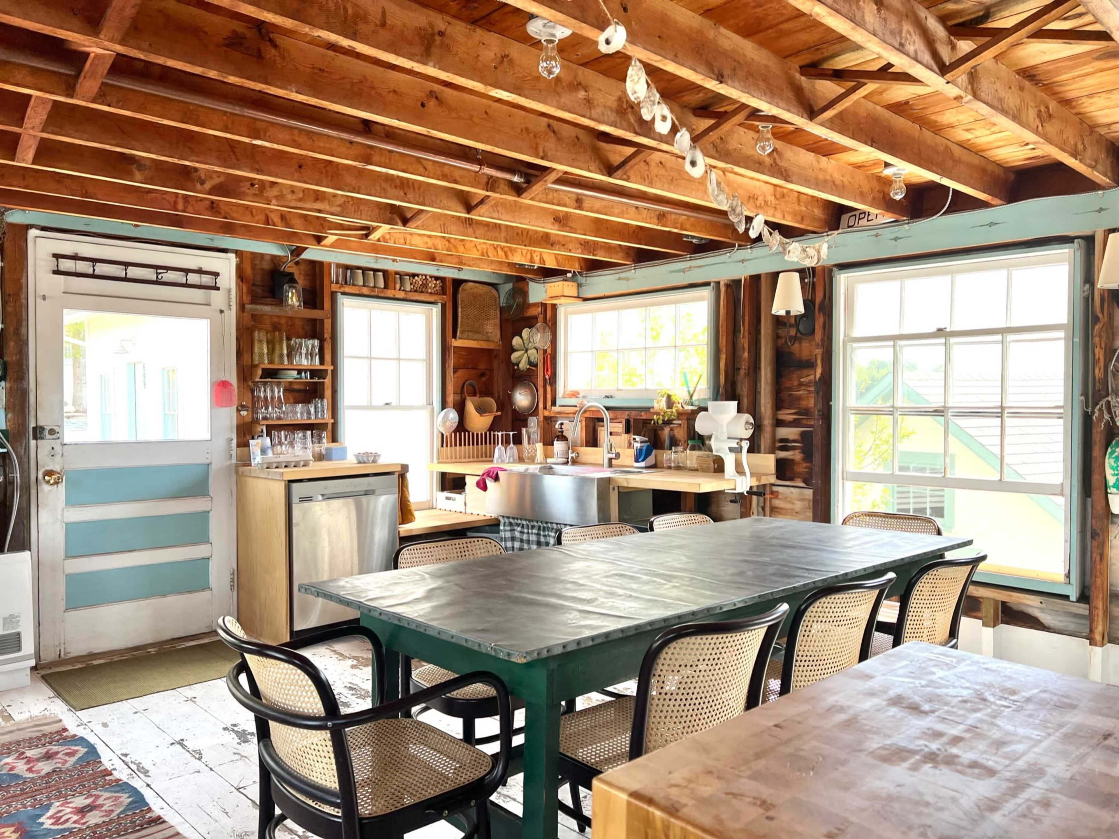 The interior features a rustic dining area with a large green table surrounded by woven chairs, a kitchen area in the background, and large windows allowing natural light to brighten the space.