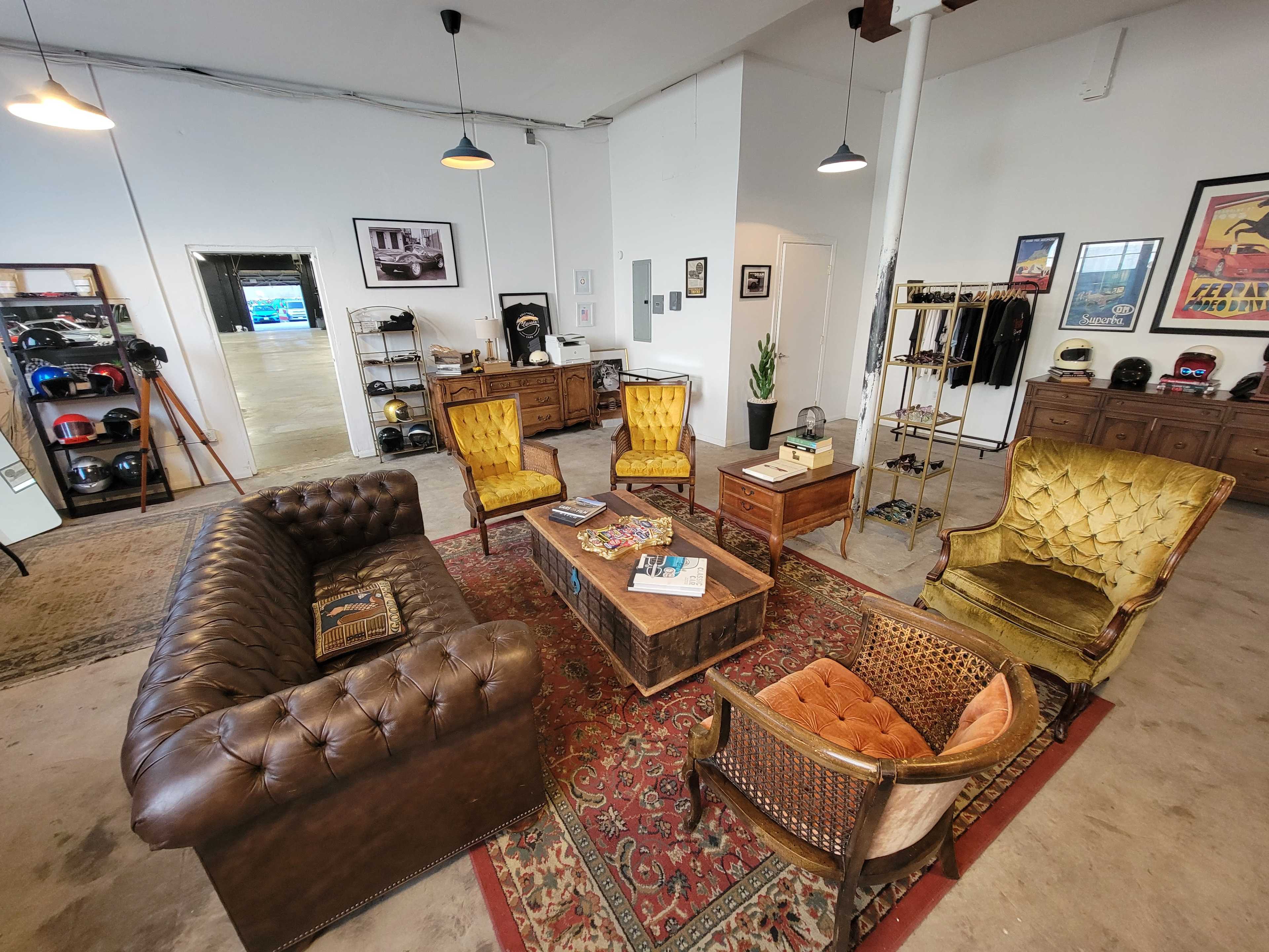 The image shows a stylish, modern office space with a leather sofa, vintage chairs, a wooden coffee table, and various decorative items and helmets on display.