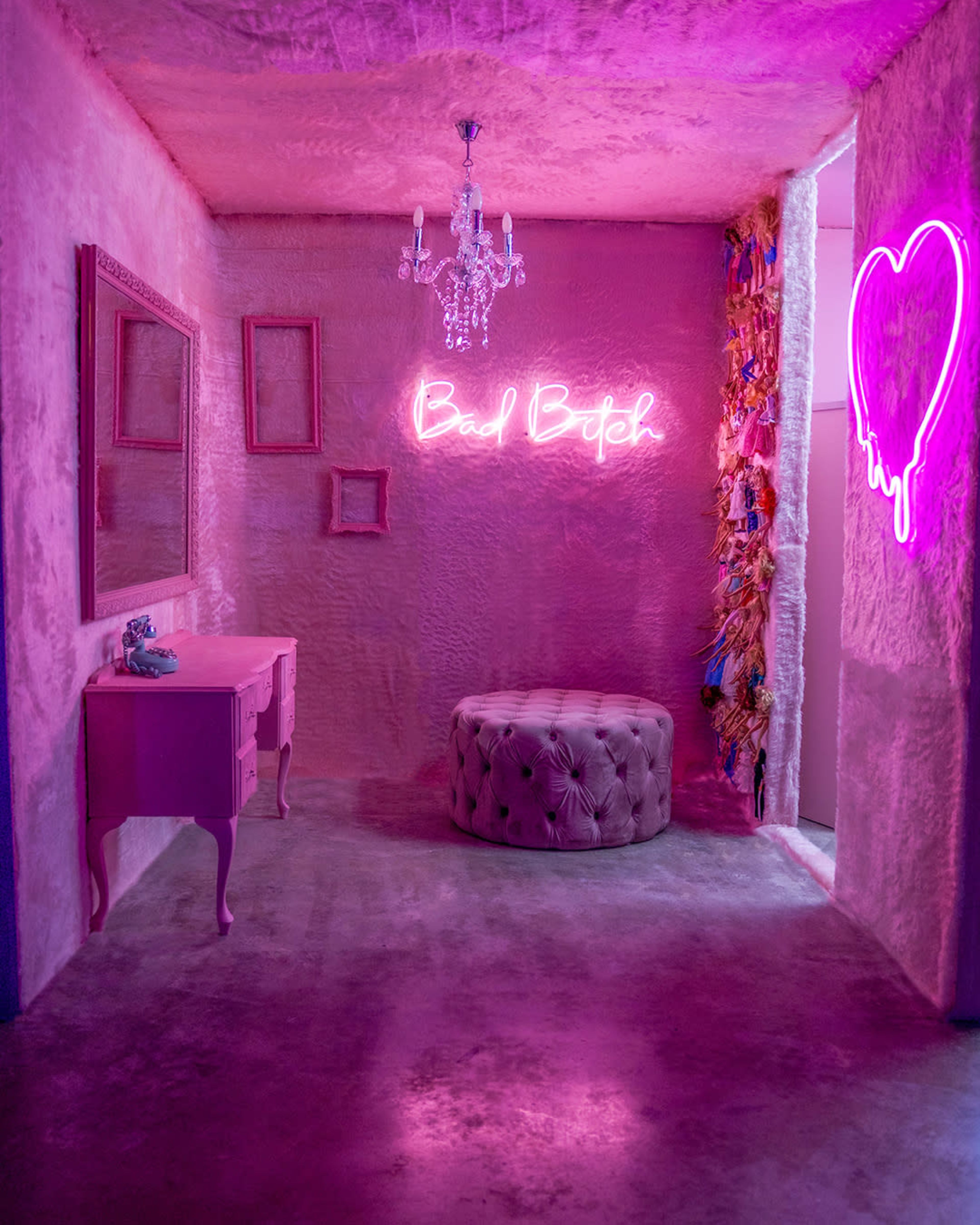 A pink-themed room with a chandelier, a mirror, a tufted ottoman, and neon signs reading "Bad Bitch" and a heart.