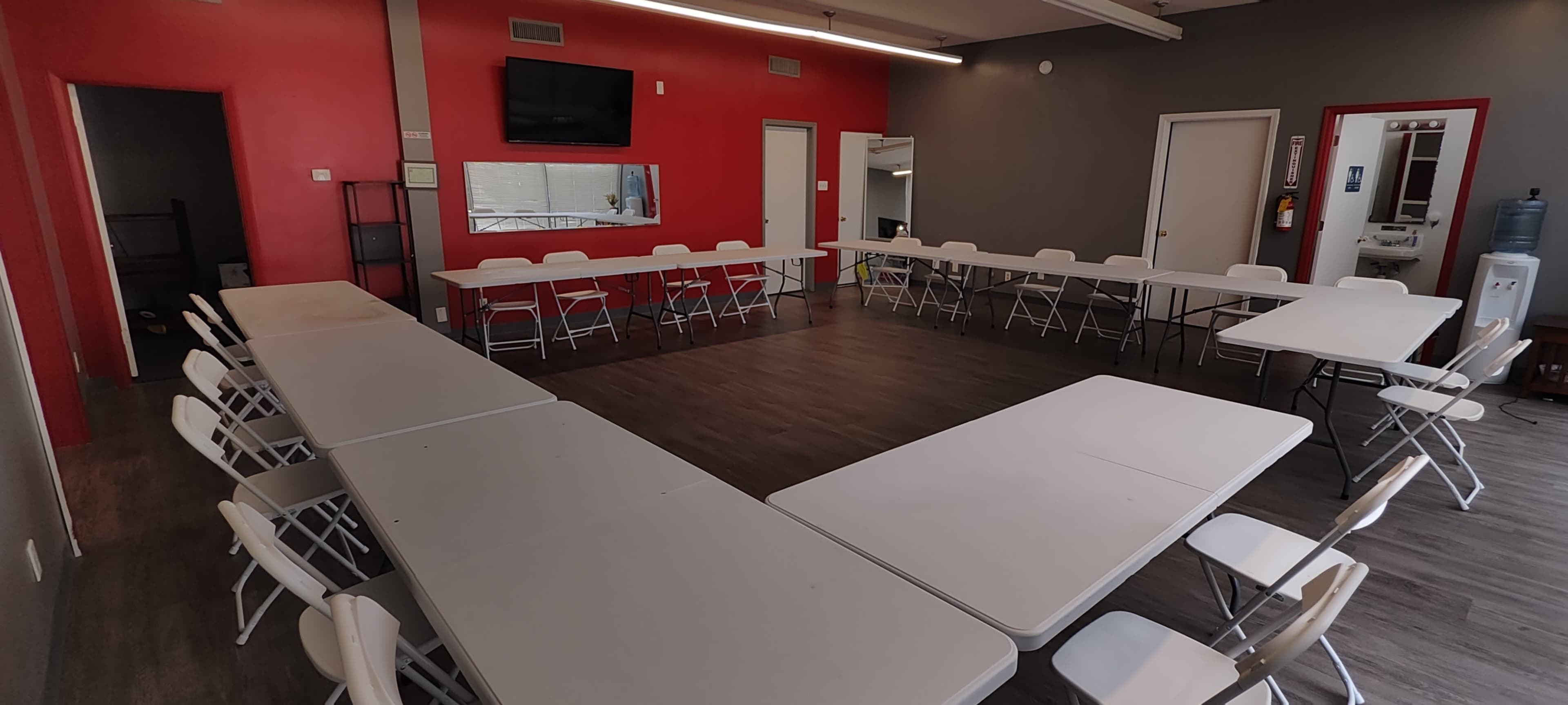 The image shows a meeting room arranged in a U-shape with multiple folding tables and chairs against a backdrop of gray and red walls, featuring a television mounted on one wall.