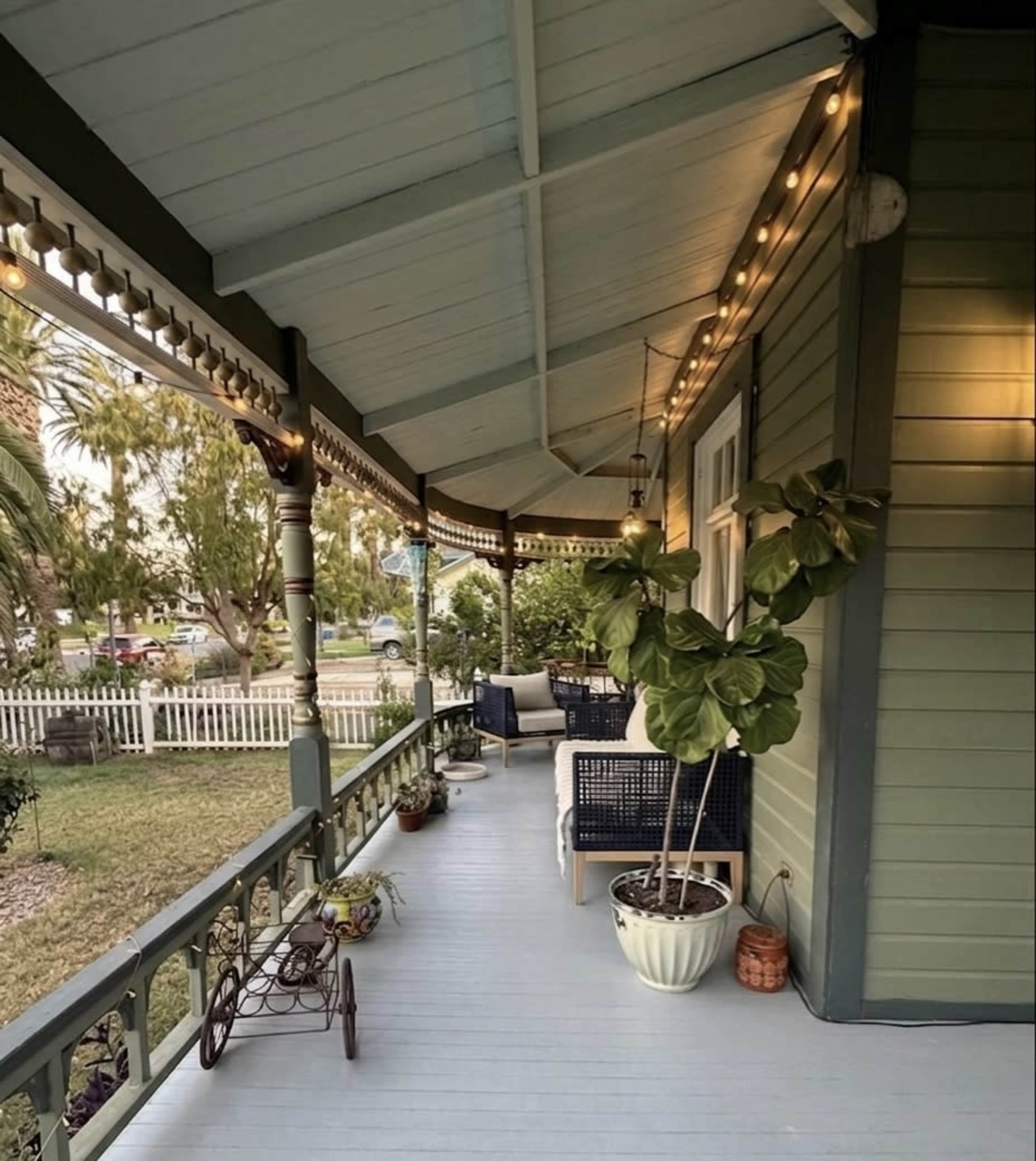 The image shows a covered porch with a wooden floor, potted plants, and string lights illuminating the space.