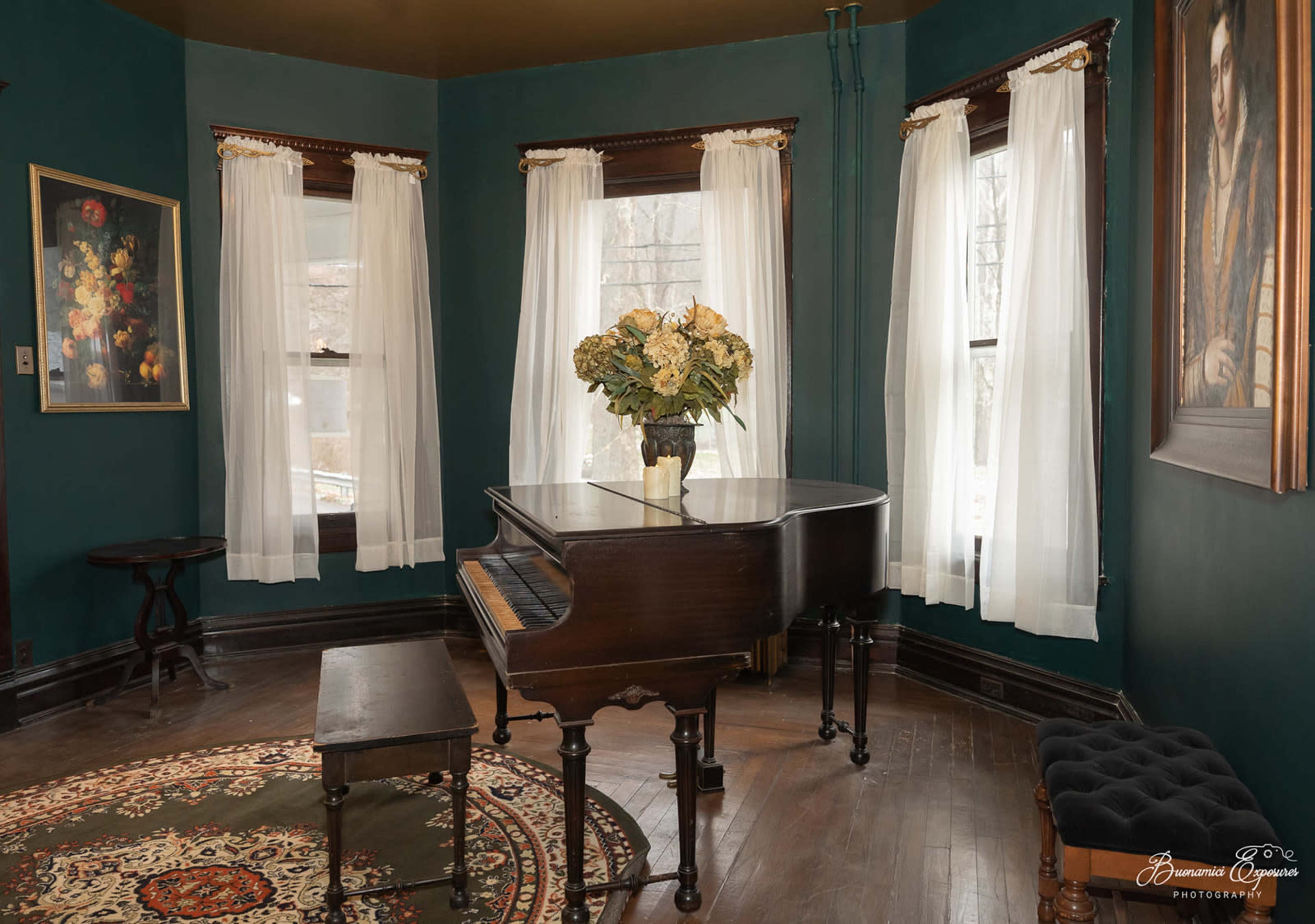 A grand piano with a floral arrangement sits in a room with large windows, dark wood trim, and a patterned rug.