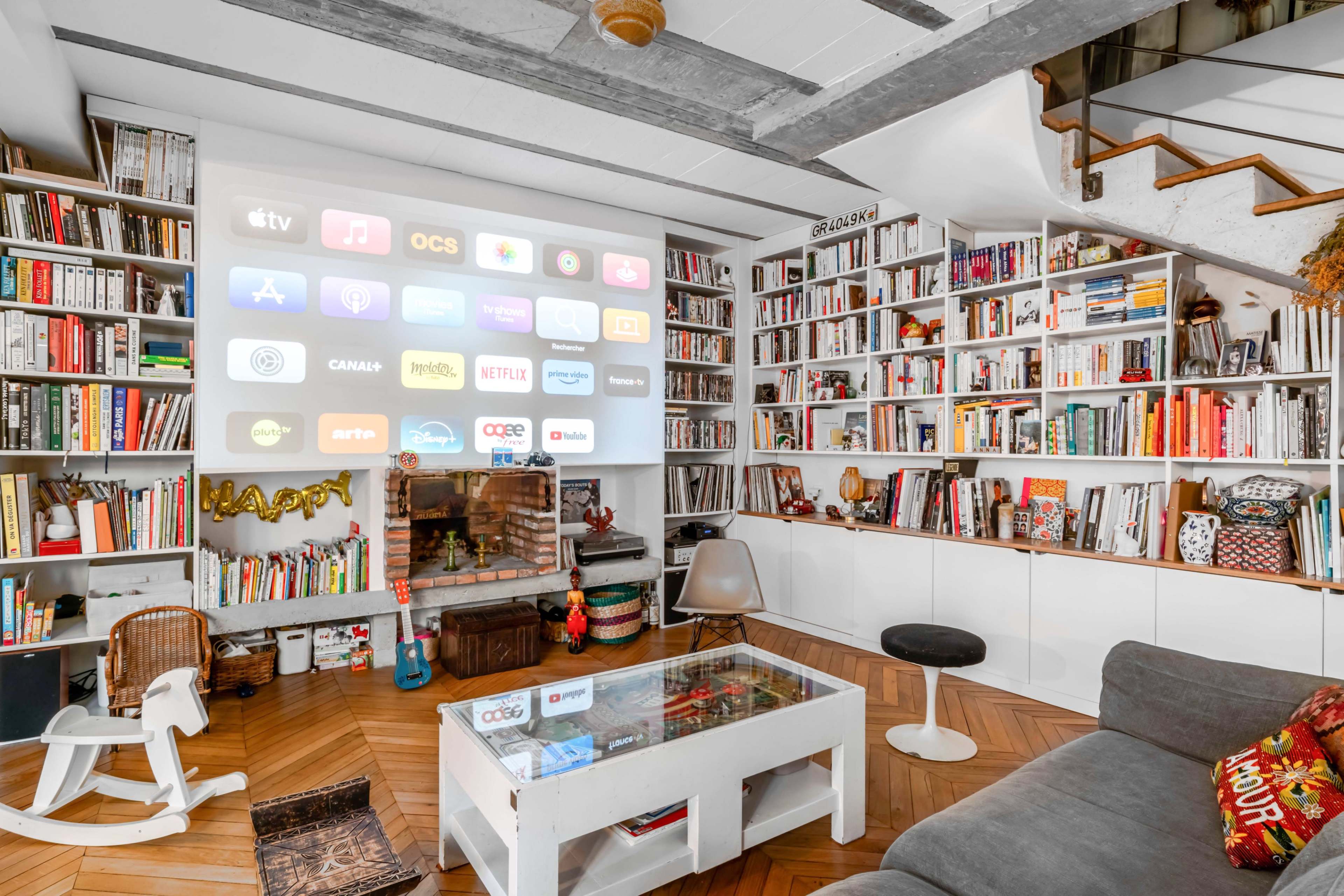 A cozy living room features a large bookshelf filled with books, a media projector displaying various streaming services on the wall, and a white coffee table in front of a comfortable seating area.