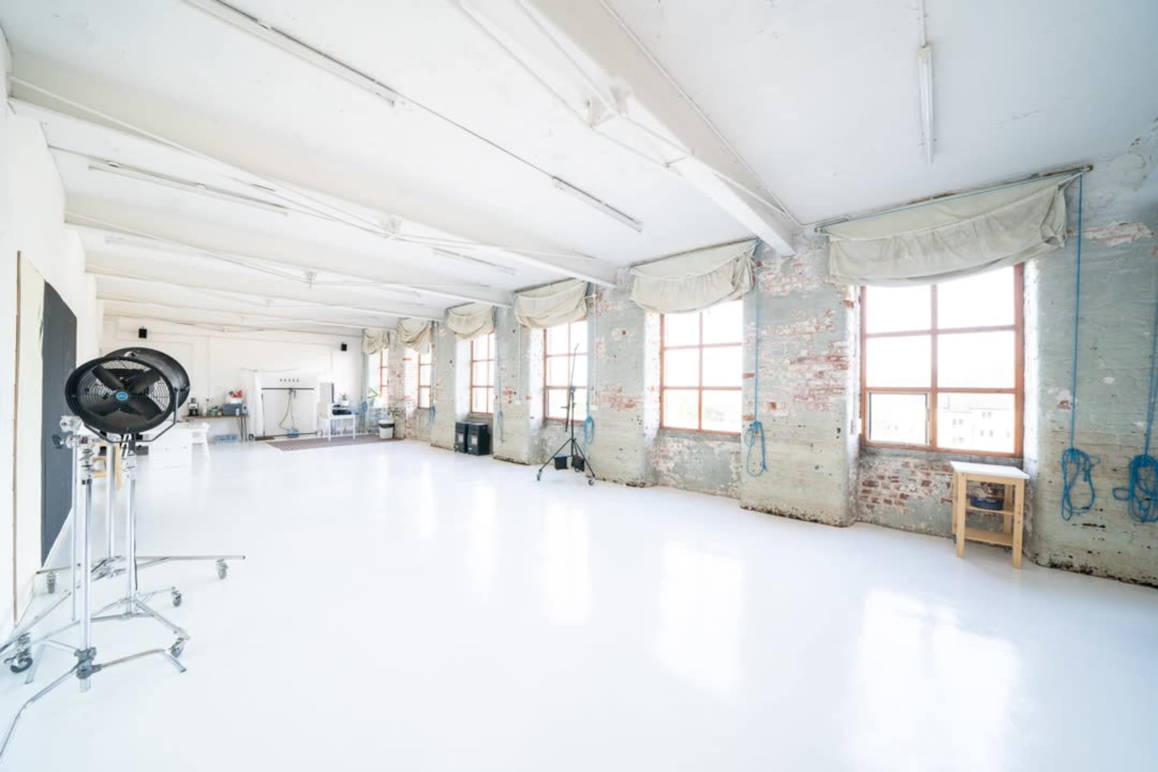 A spacious, well-lit studio with large windows and a polished white floor, featuring exposed brick walls and minimal furnishings.