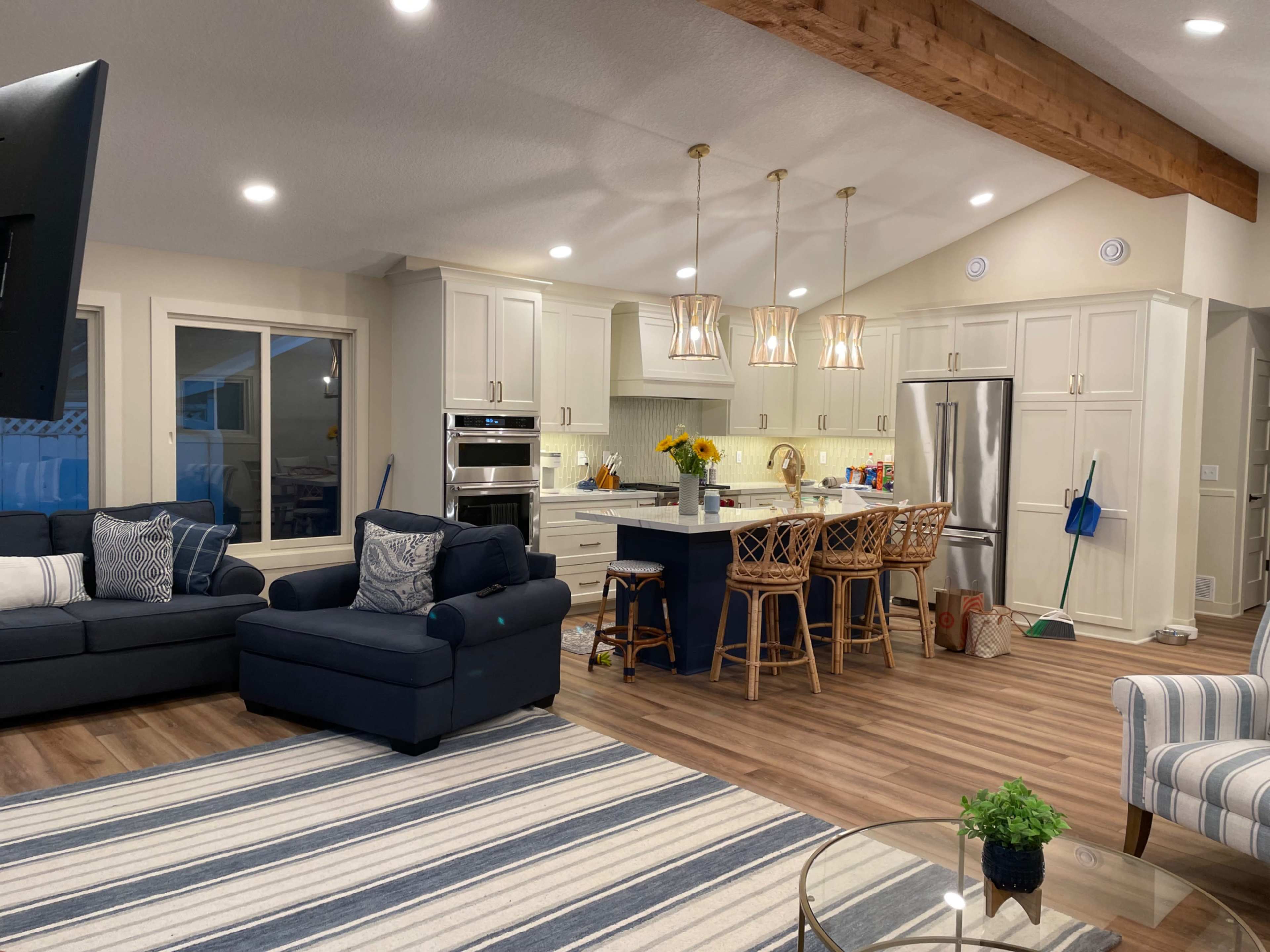 A modern kitchen and living area with light-colored cabinetry, four bar stools at the island, and a sectional sofa with a striped rug.