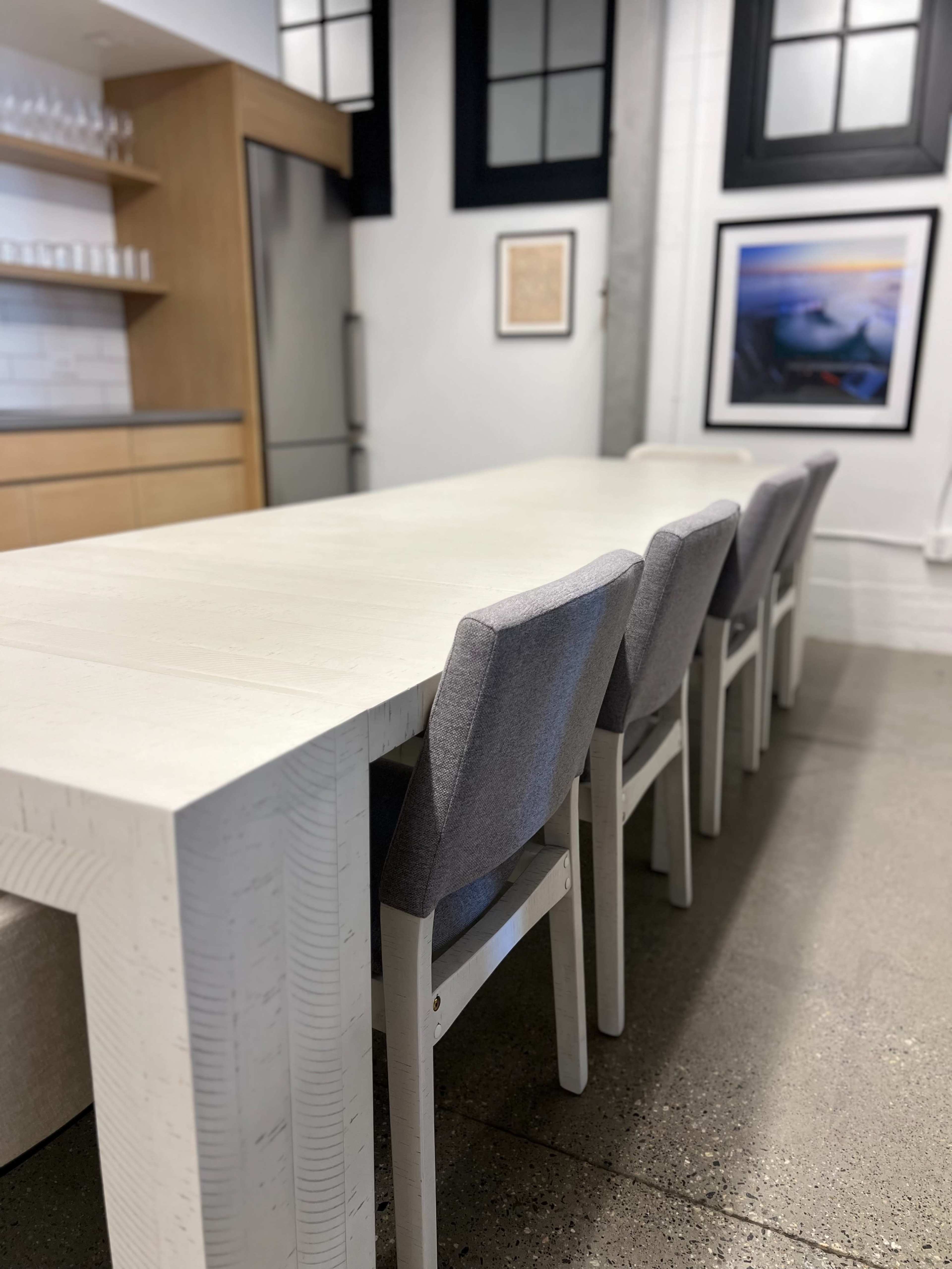 A modern kitchen features a long white table with gray chairs, set against a backdrop of large windows and contemporary cabinetry.
