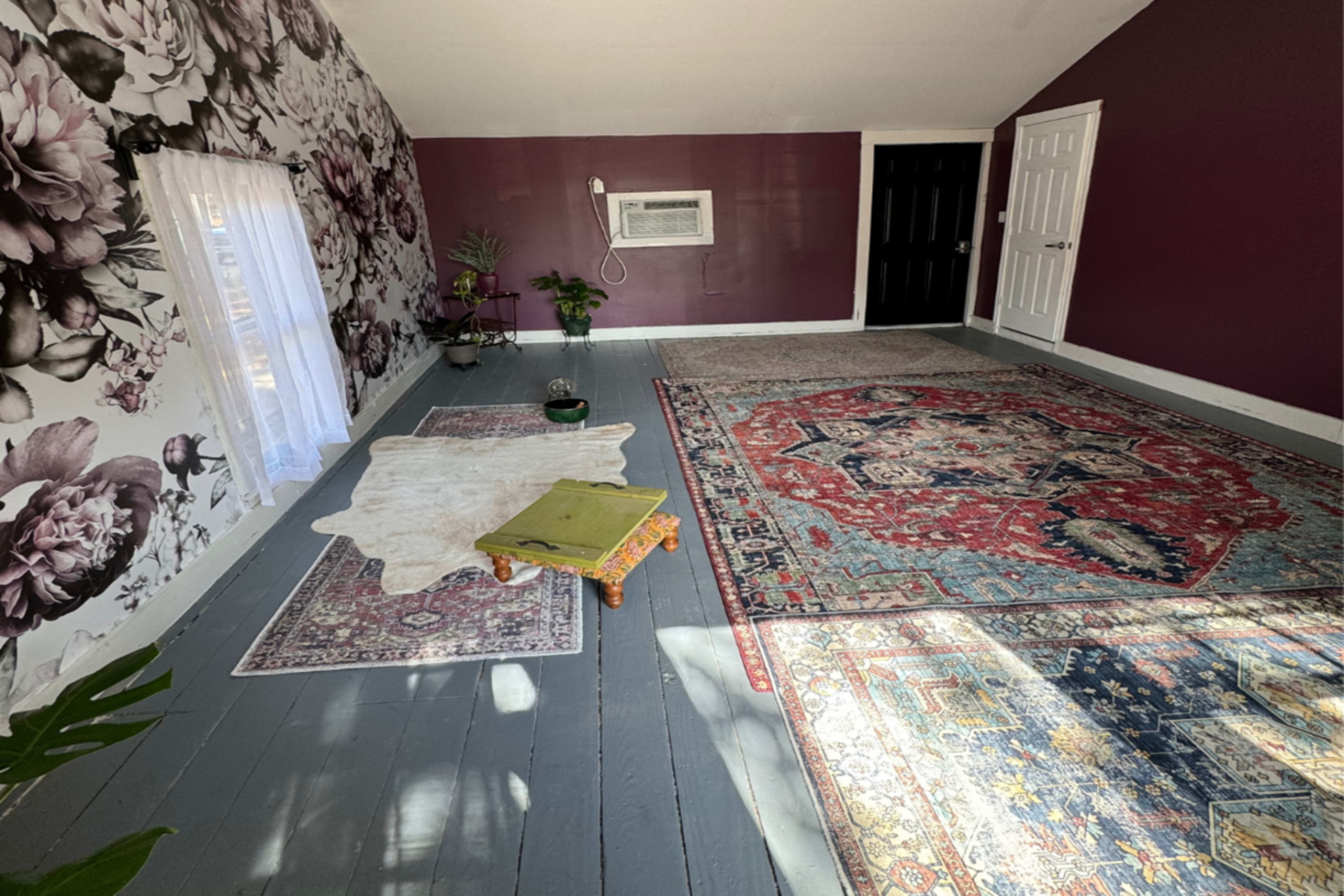 The image shows a spacious attic room with floral wallpaper, two area rugs, a small table, and a window partially covered by a sheer curtain.