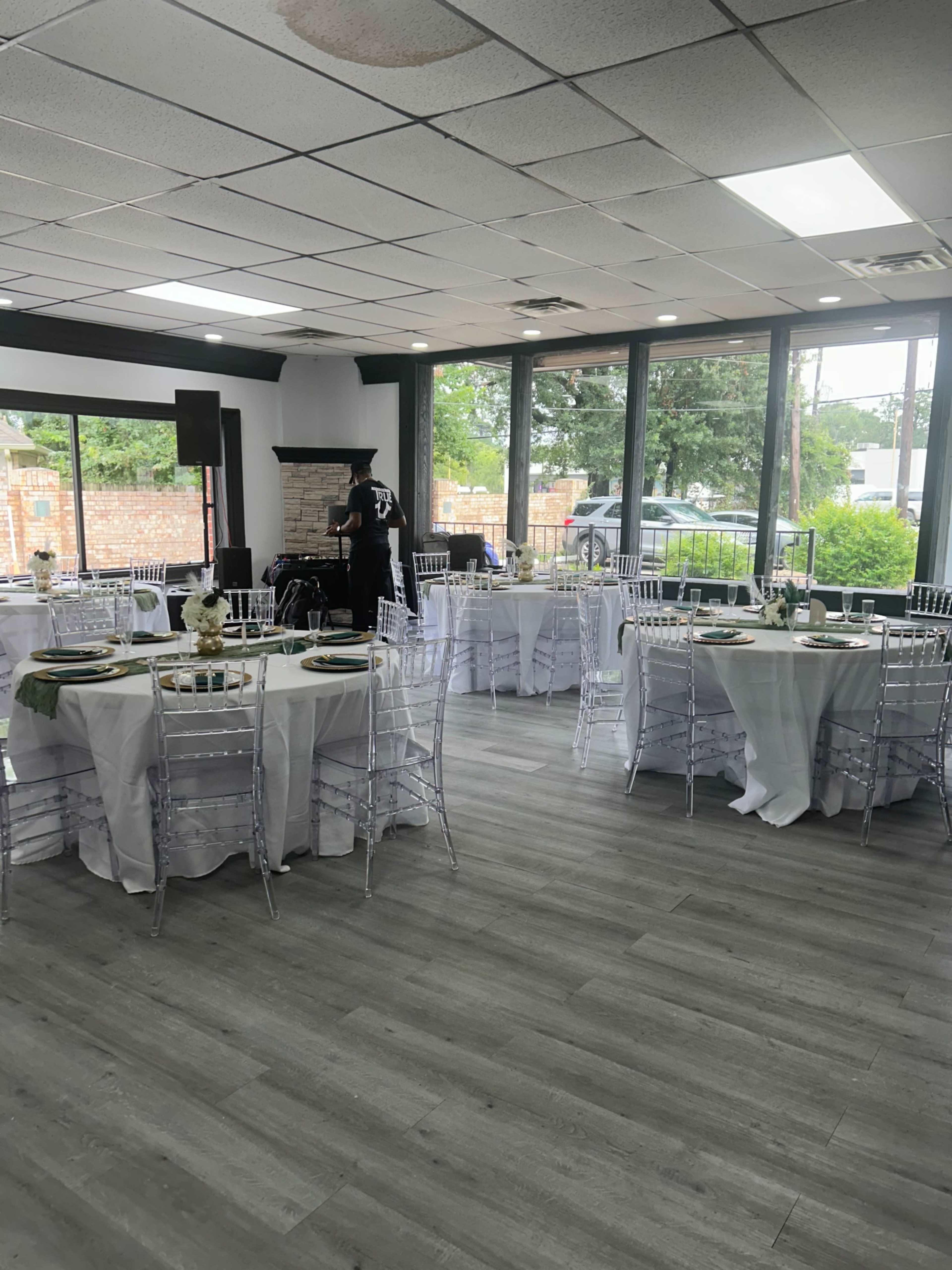 The image shows a decorated event space with multiple round tables set for a gathering, featuring white tablecloths and clear chairs, and a person arranging items in the background.