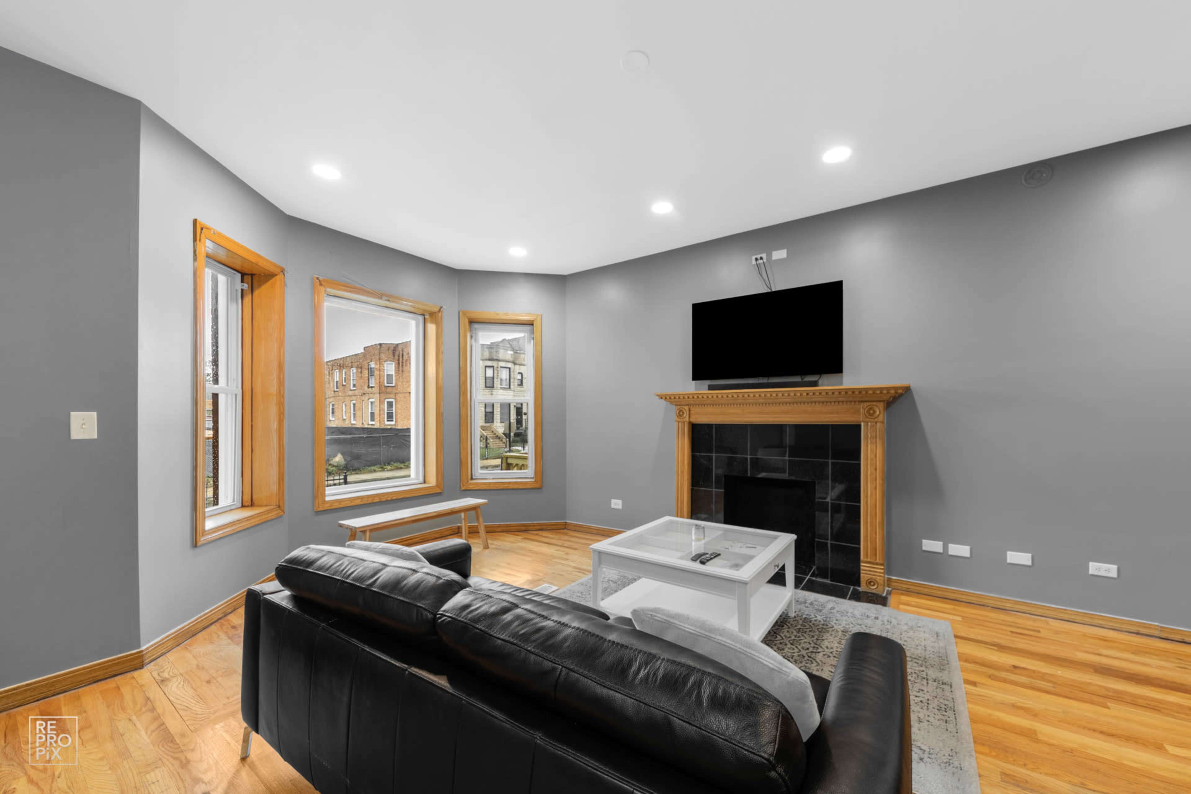 The room features a black leather couch facing a flat-screen TV mounted above a fireplace, with large windows providing a view of the street outside.