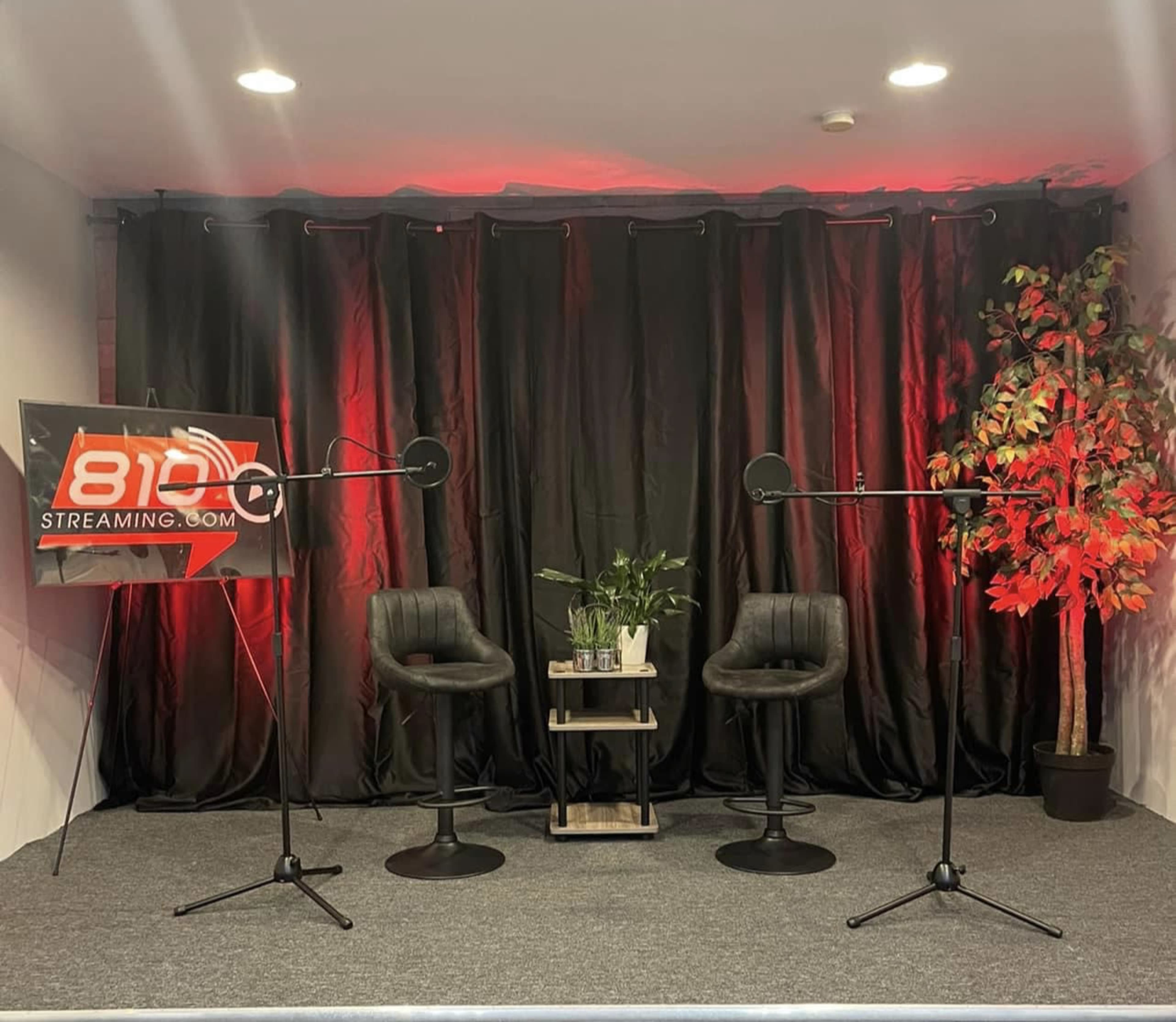 A small stage setup with two black chairs, two microphone stands, a potted plant, and a sign for "810 Streaming," all against a dark curtain backdrop.