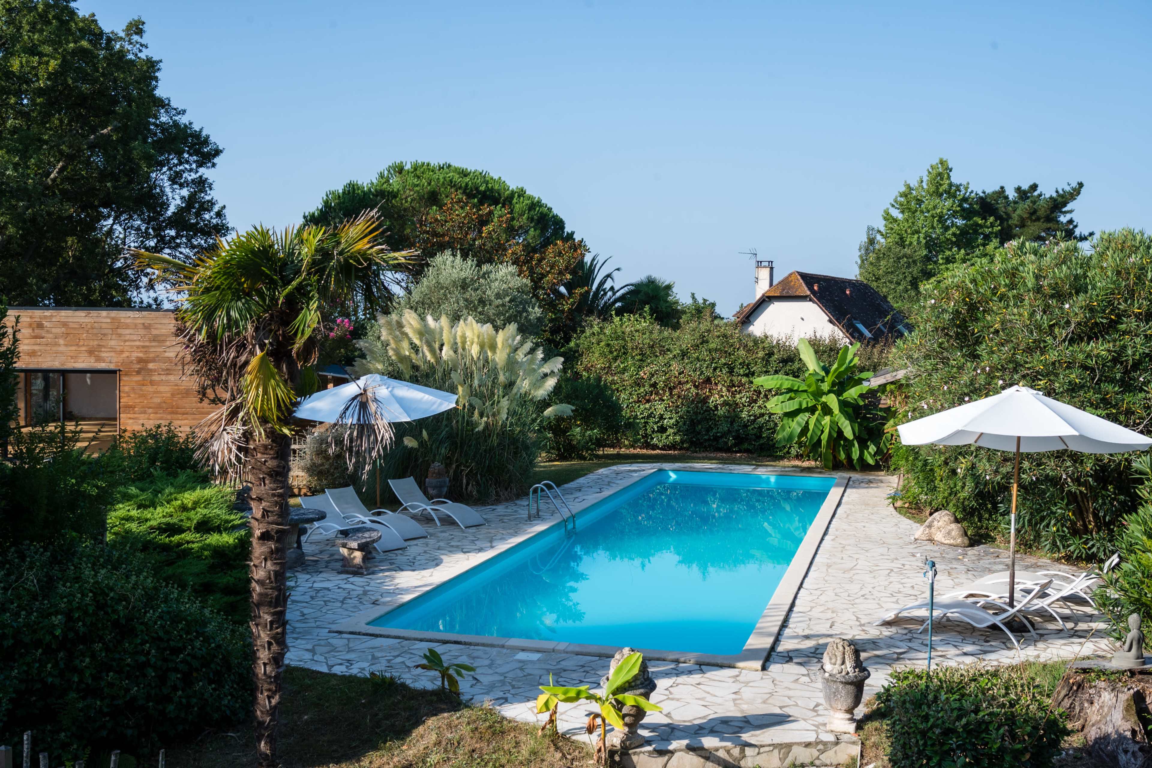 A clear blue swimming pool is surrounded by lush greenery and sun loungers under white umbrellas.