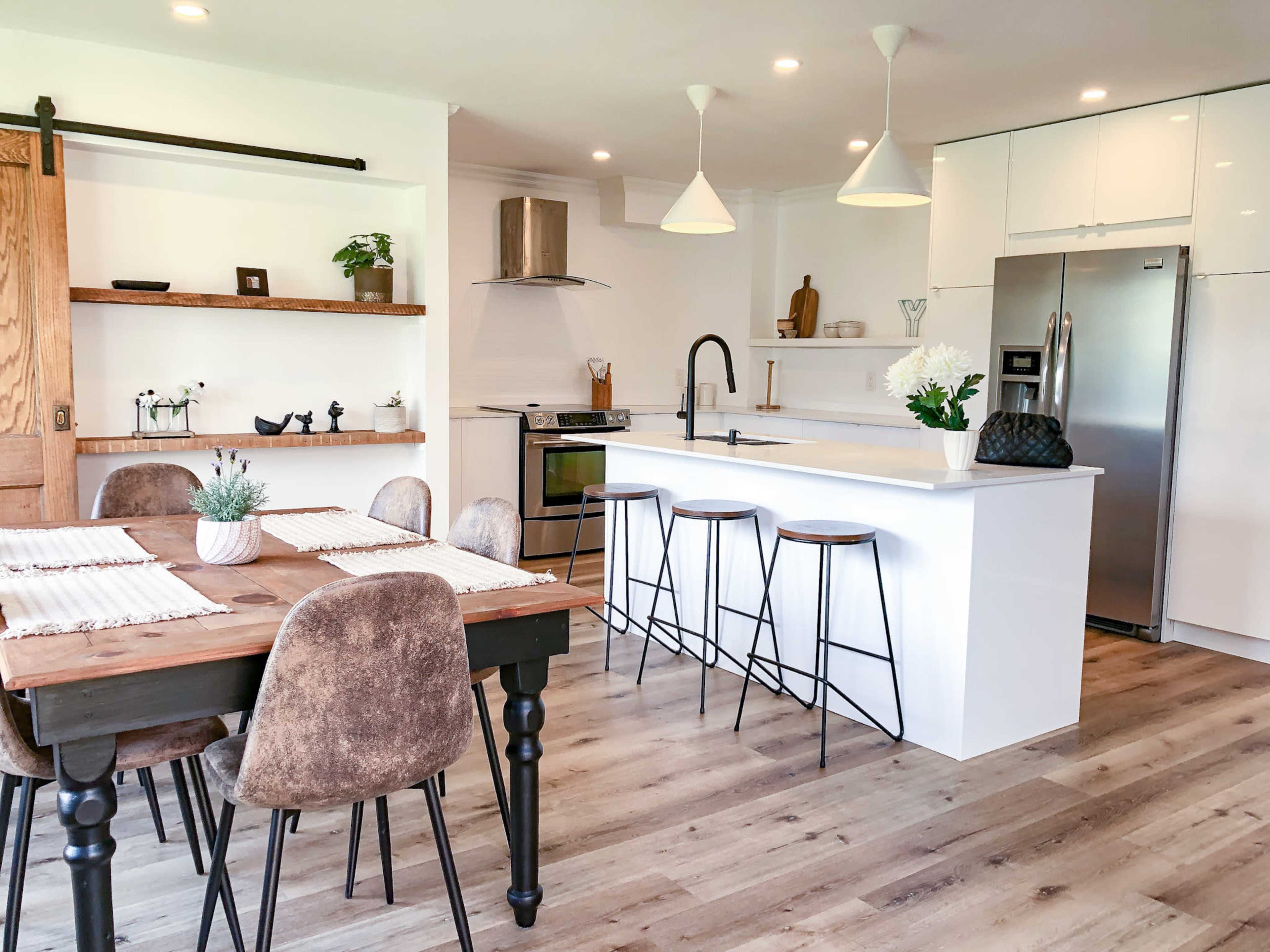 A modern kitchen and dining area featuring a wooden table with upholstered chairs, a sleek white countertop with bar stools, and open shelving with decor.