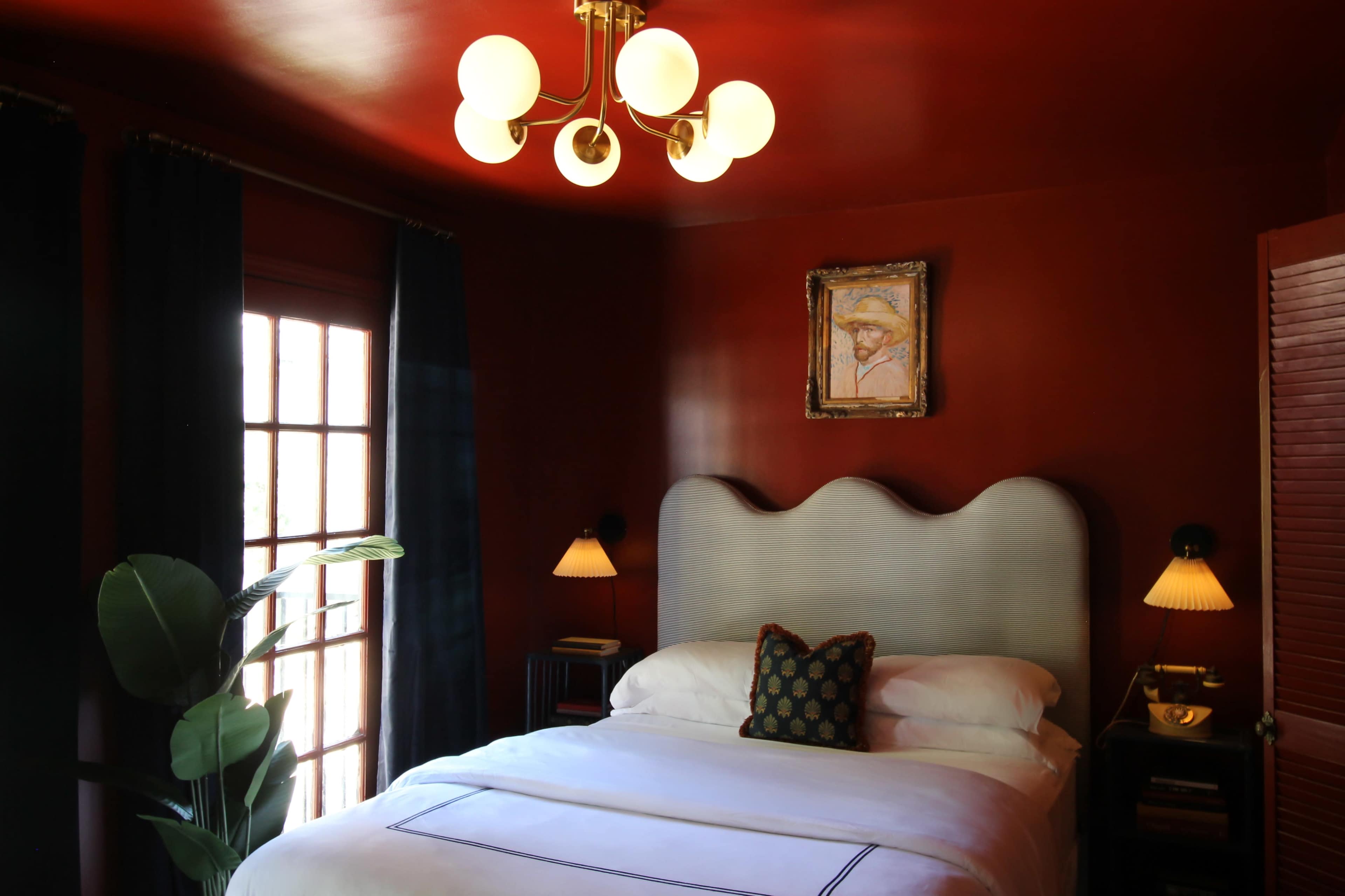 The image shows a bedroom with a red-painted wall, a bed with a curved headboard, and a chandelier above, along with a portrait on the wall and plants by the window.