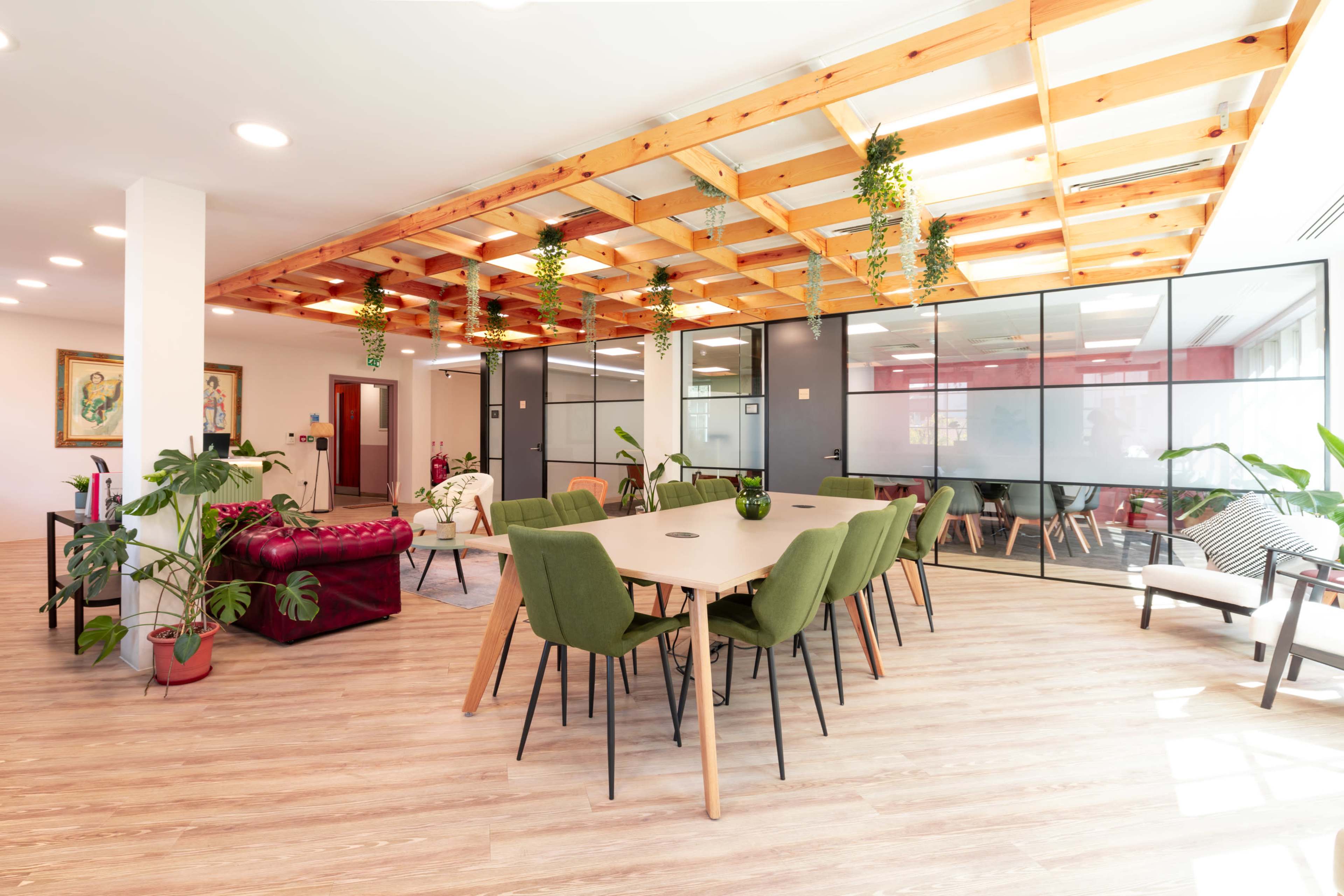 The image shows a modern office space with a dining table surrounded by green chairs, a lounge area with a burgundy couch, and glass meeting rooms.