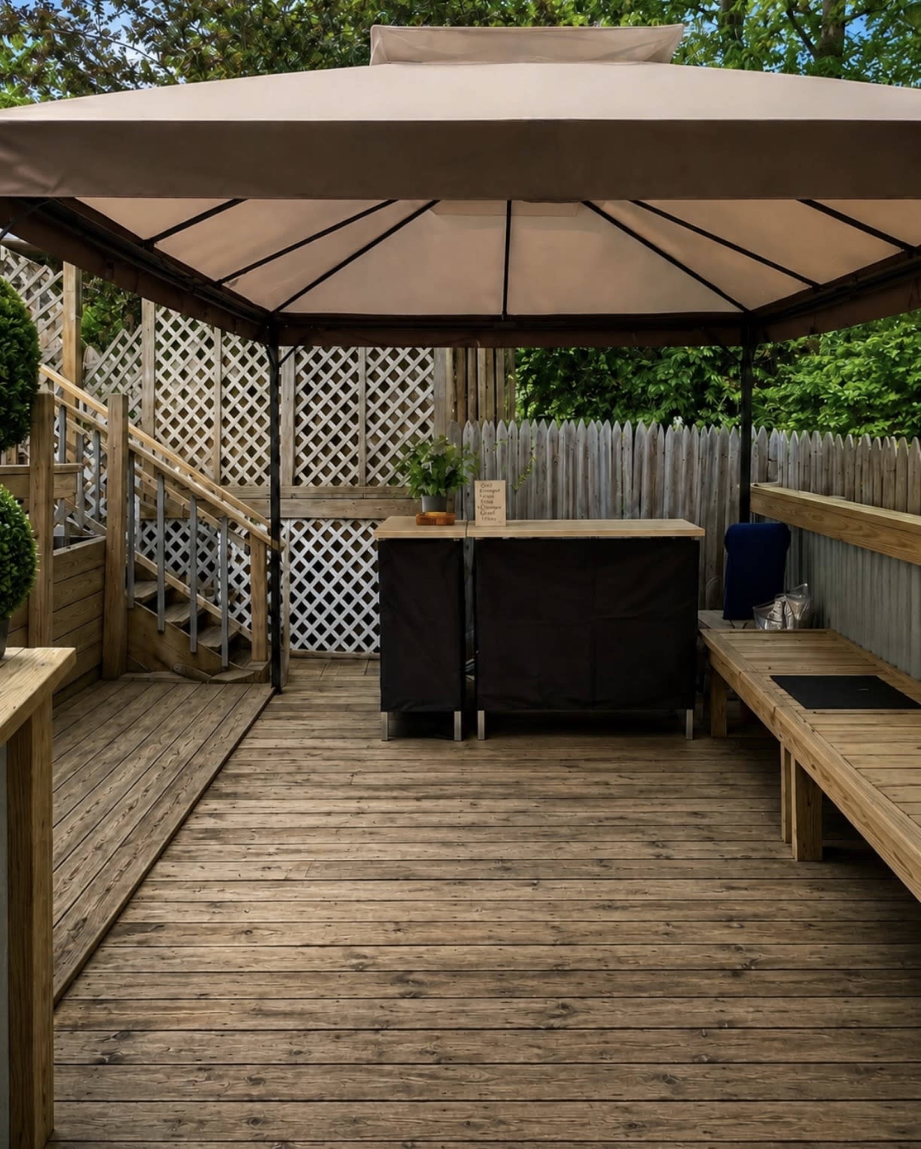 The image shows a wooden patio with a canopy, a bar area, and a staircase leading to a fenced garden.