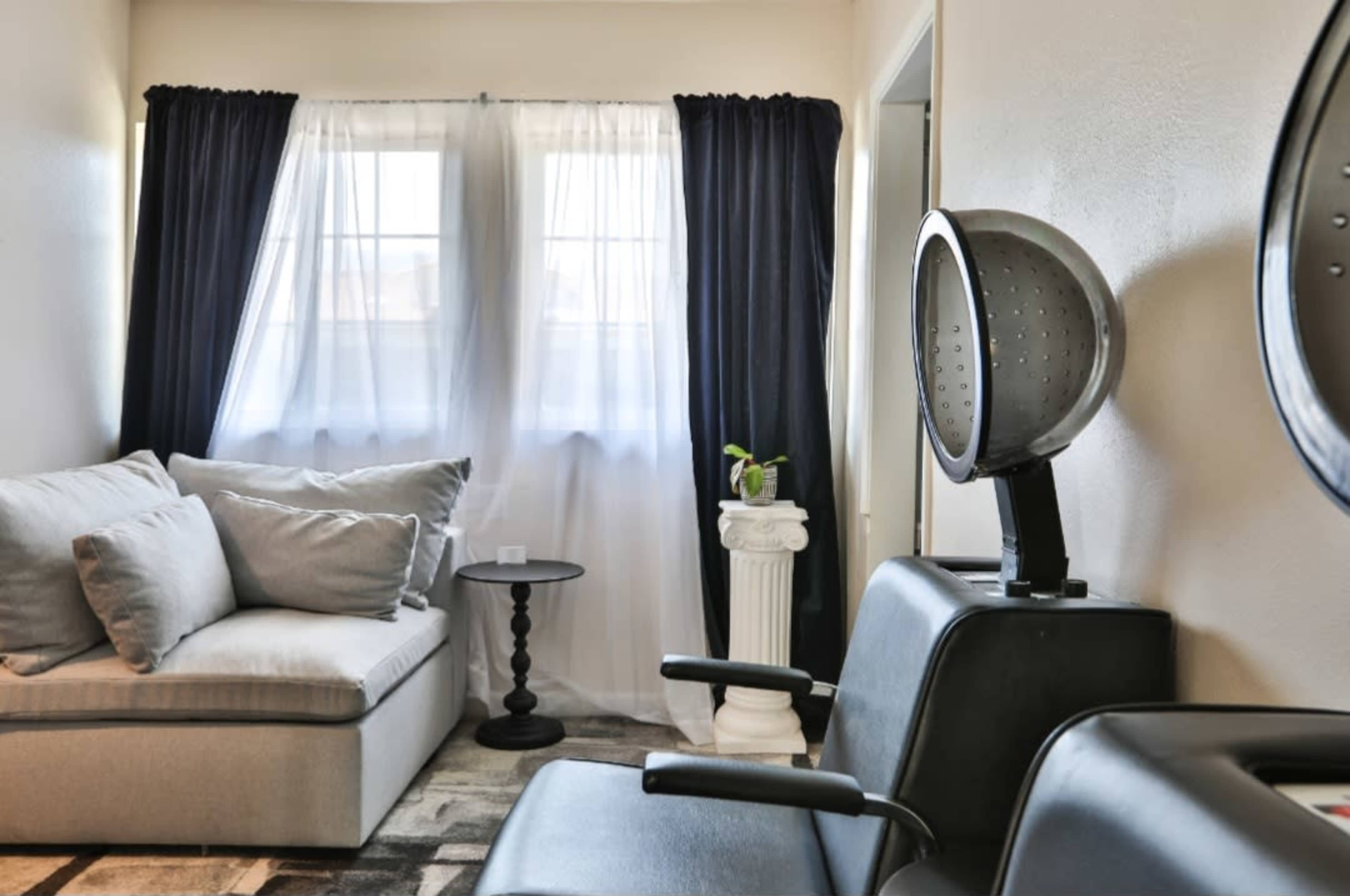 A cozy salon waiting area features a gray sofa, an end table, and salon chairs with a hair dryer positioned nearby.