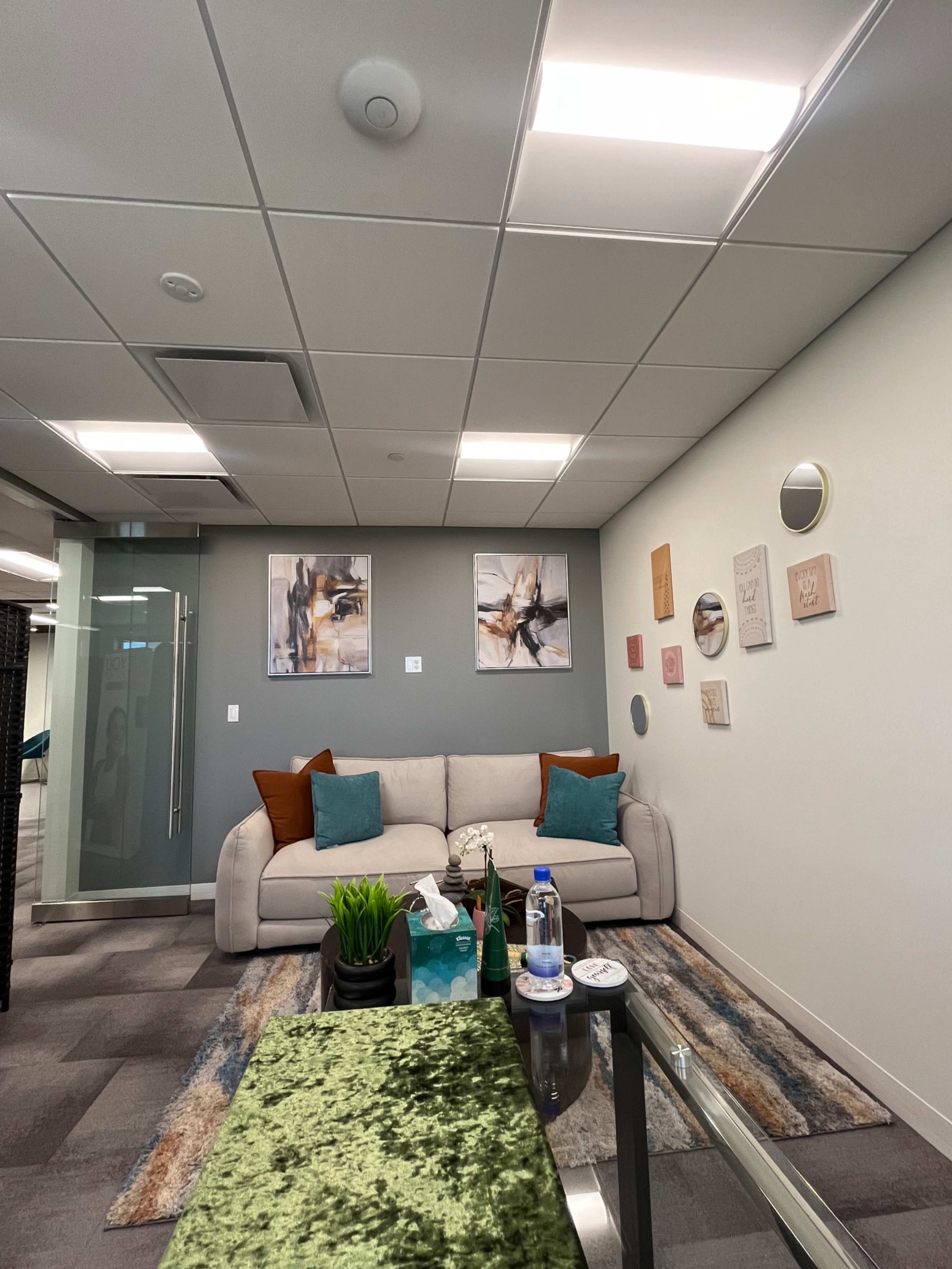 A modern office lounge area featuring a light-colored sofa with decorative pillows, a coffee table, artwork on the walls, and a plant.