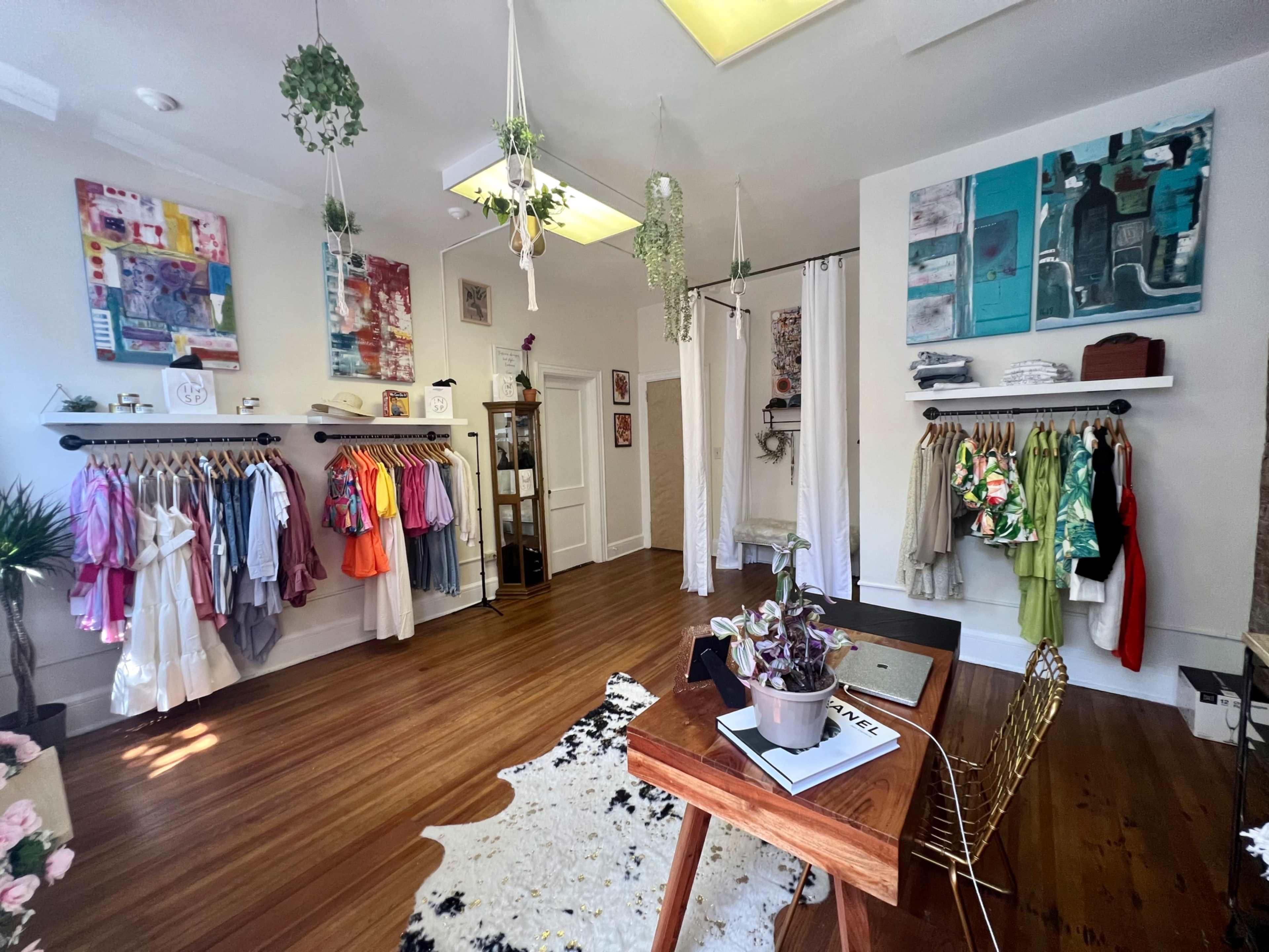 A bright boutique interior featuring clothing racks, artwork on the walls, and a small table with a plant.