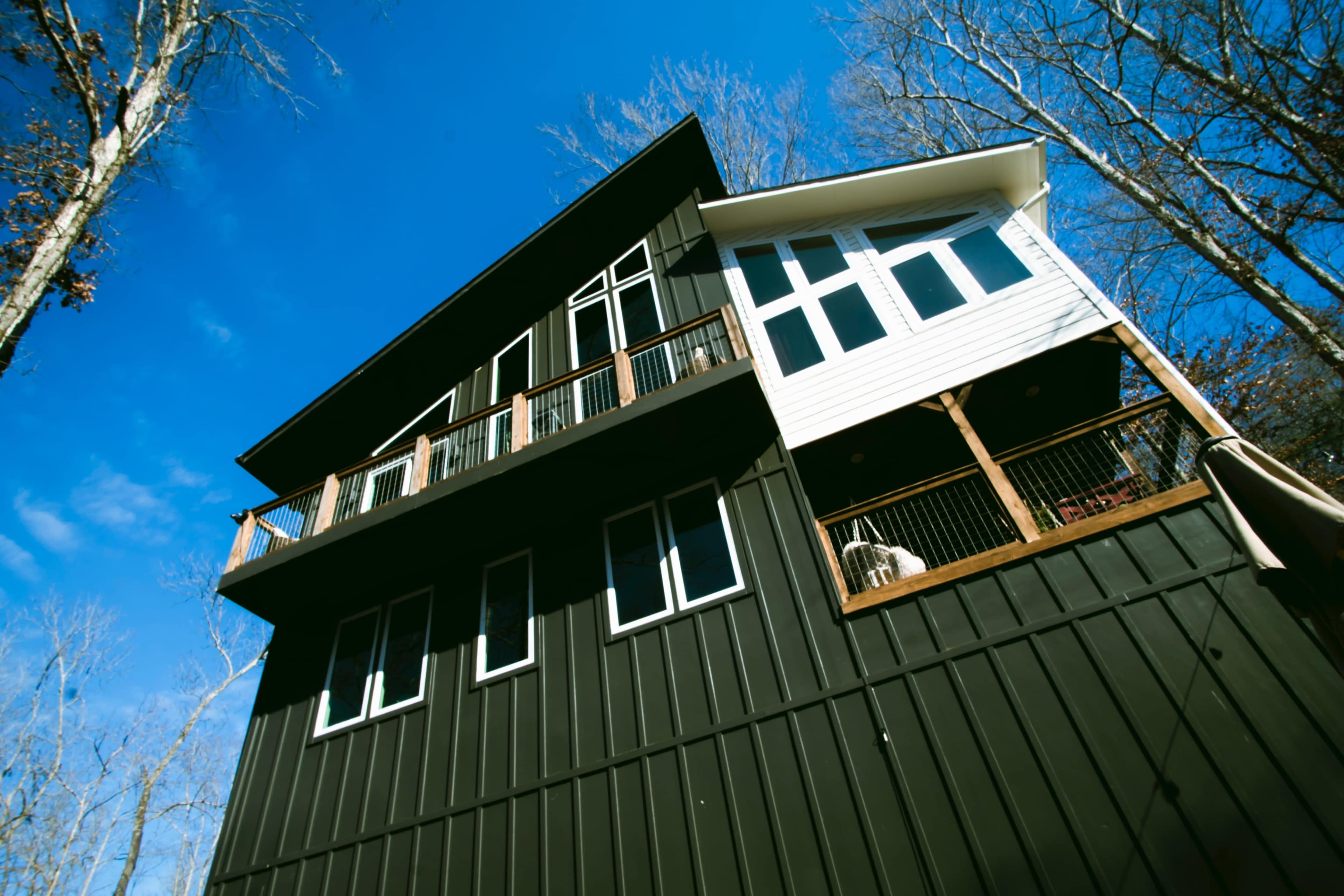The image shows a modern two-story house with a mix of dark siding and white panels, surrounded by bare trees under a clear blue sky.