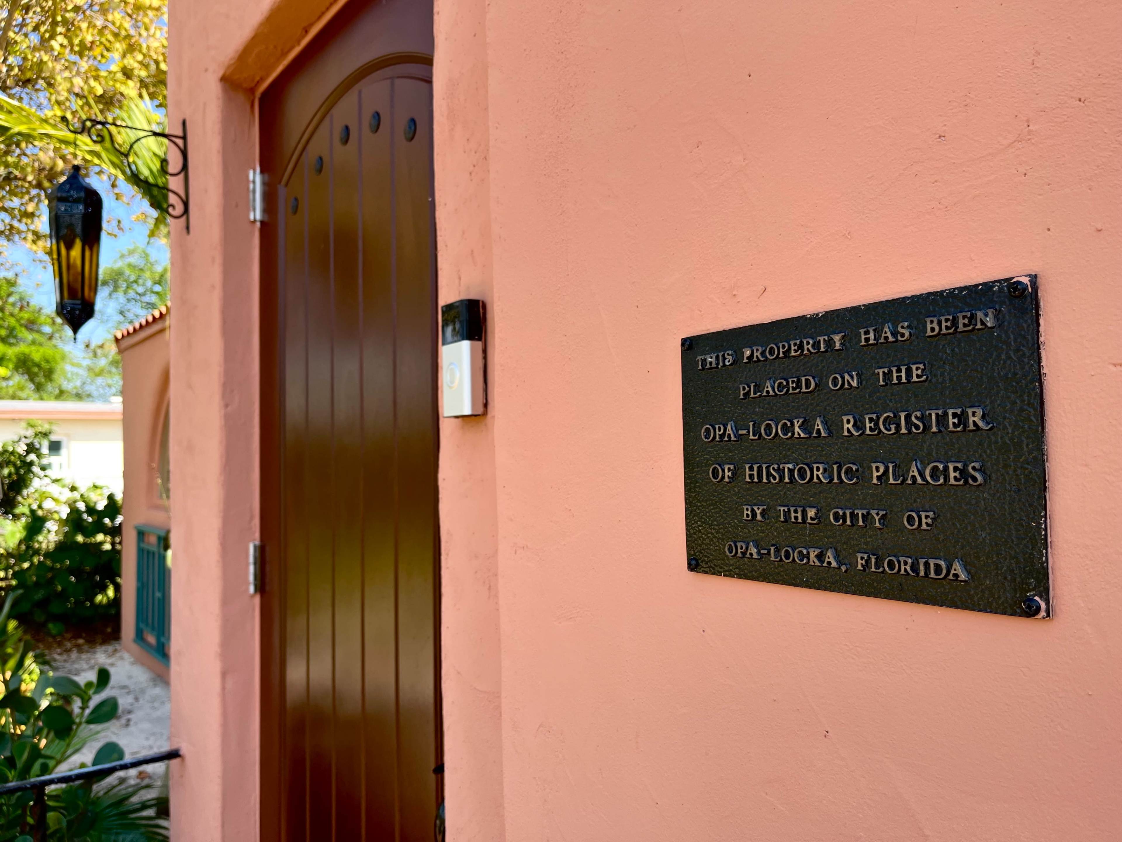 A bronze plaque on a pink wall indicates that the property has been placed on the Opa-locka Register of Historic Places by the City of Opa-locka, Florida.