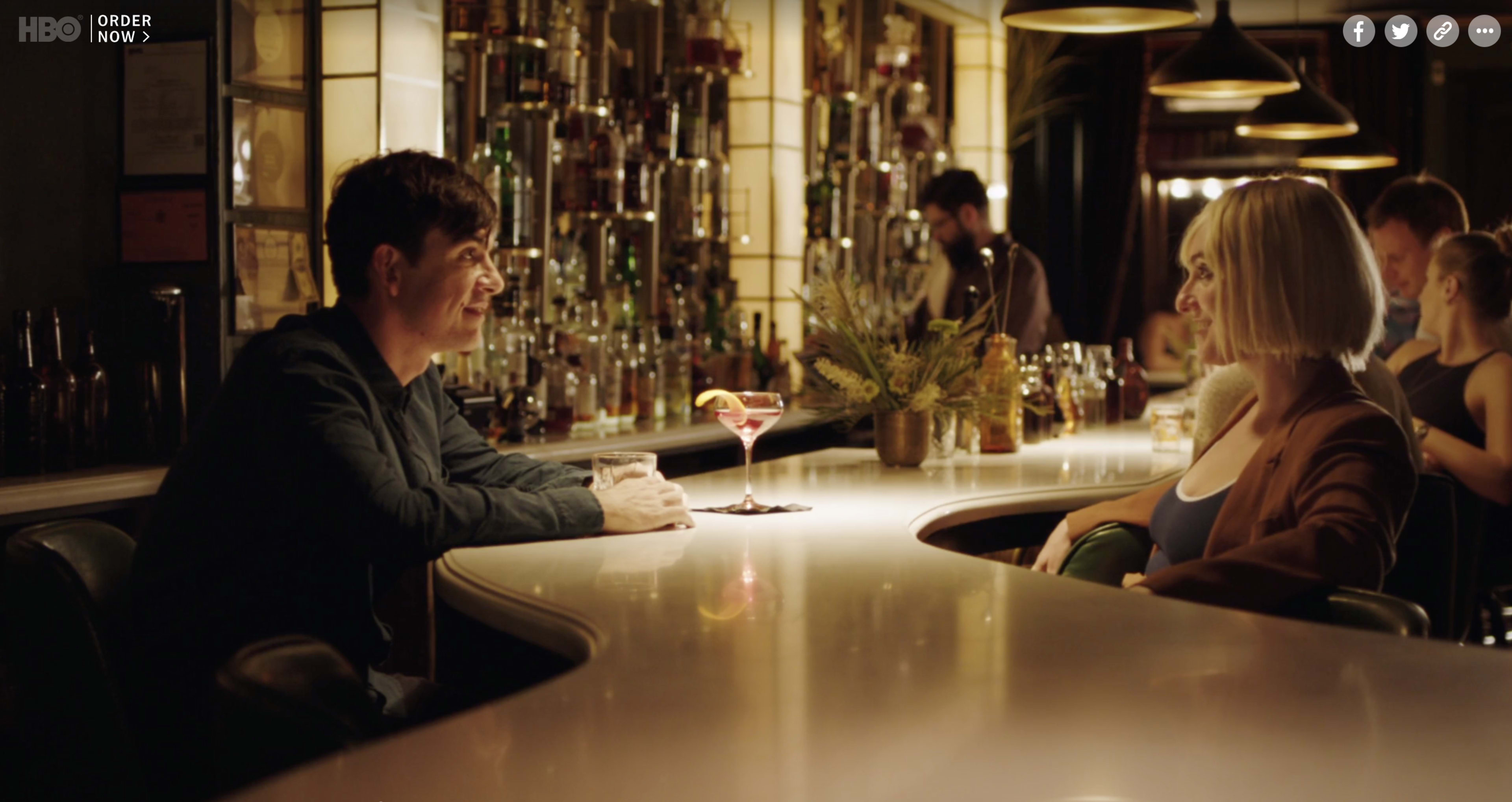 Two people are seated at a bar, with one man looking at a woman while a bartender prepares drinks in the background.