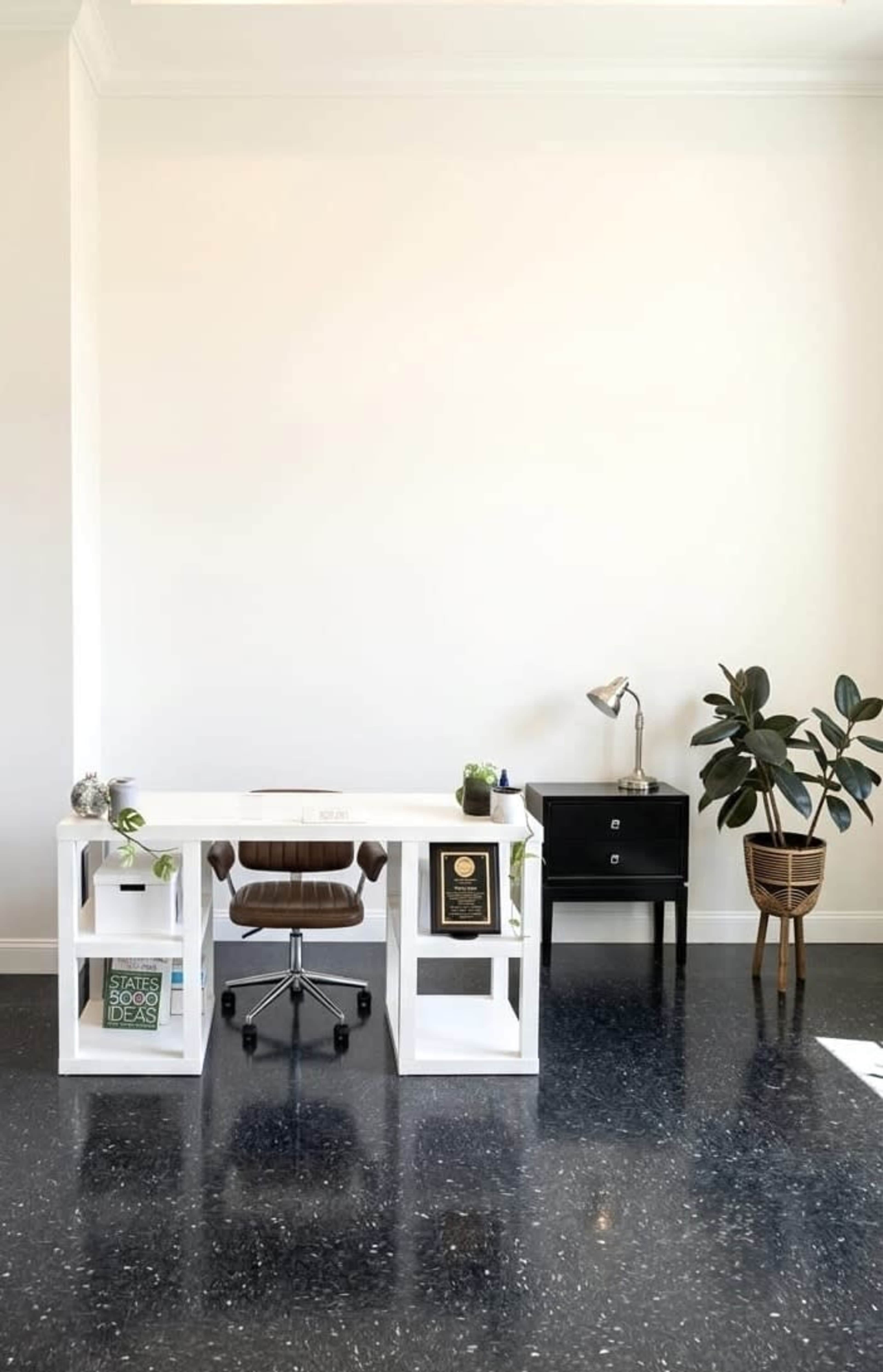 The image shows a minimalistic office space featuring a white desk with a brown chair, a small black filing cabinet, a lamp, and a potted plant.