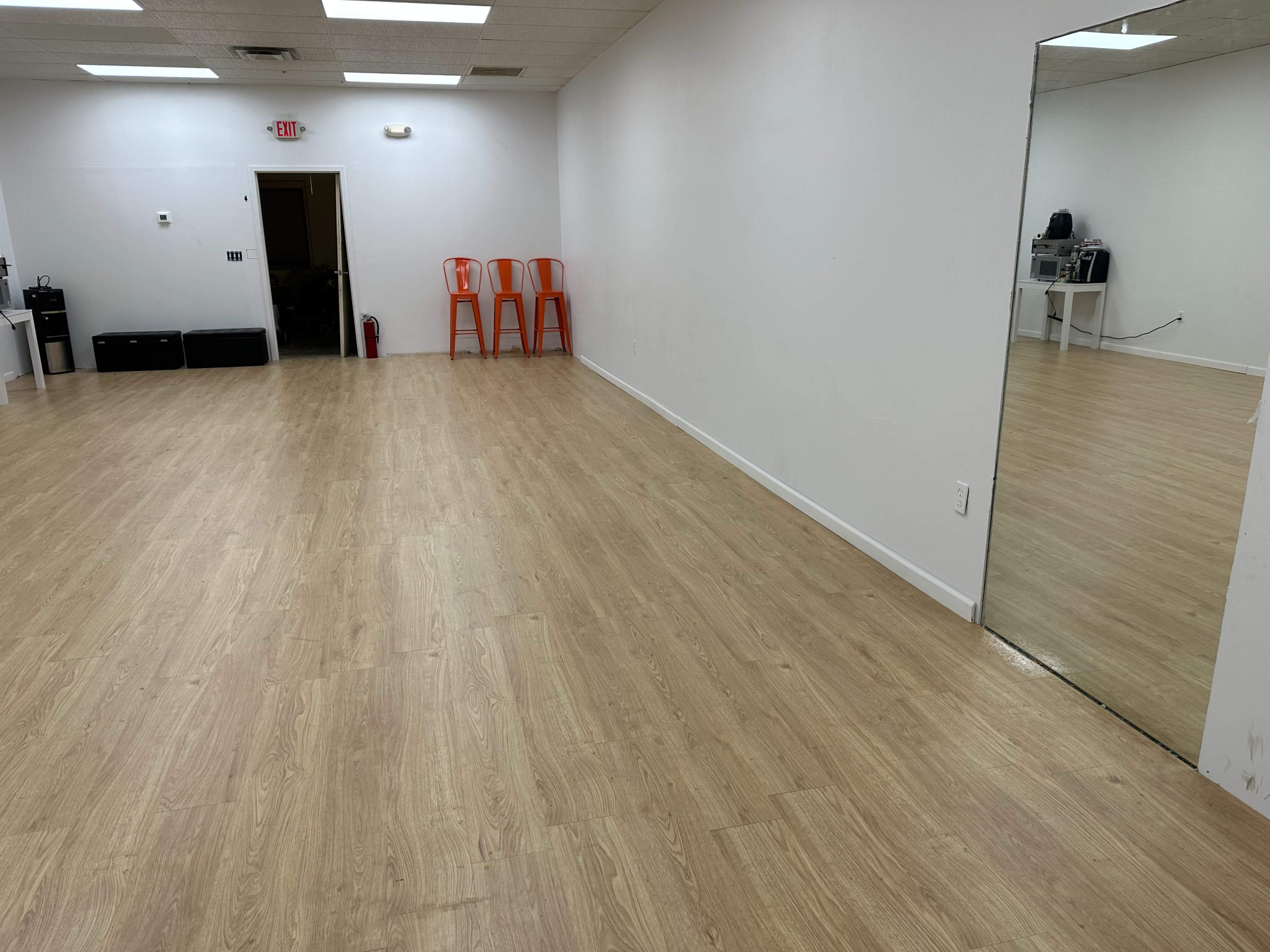 The image shows an empty room with light wooden flooring, white walls, three orange chairs stacked in the corner, and a mirror on one wall.