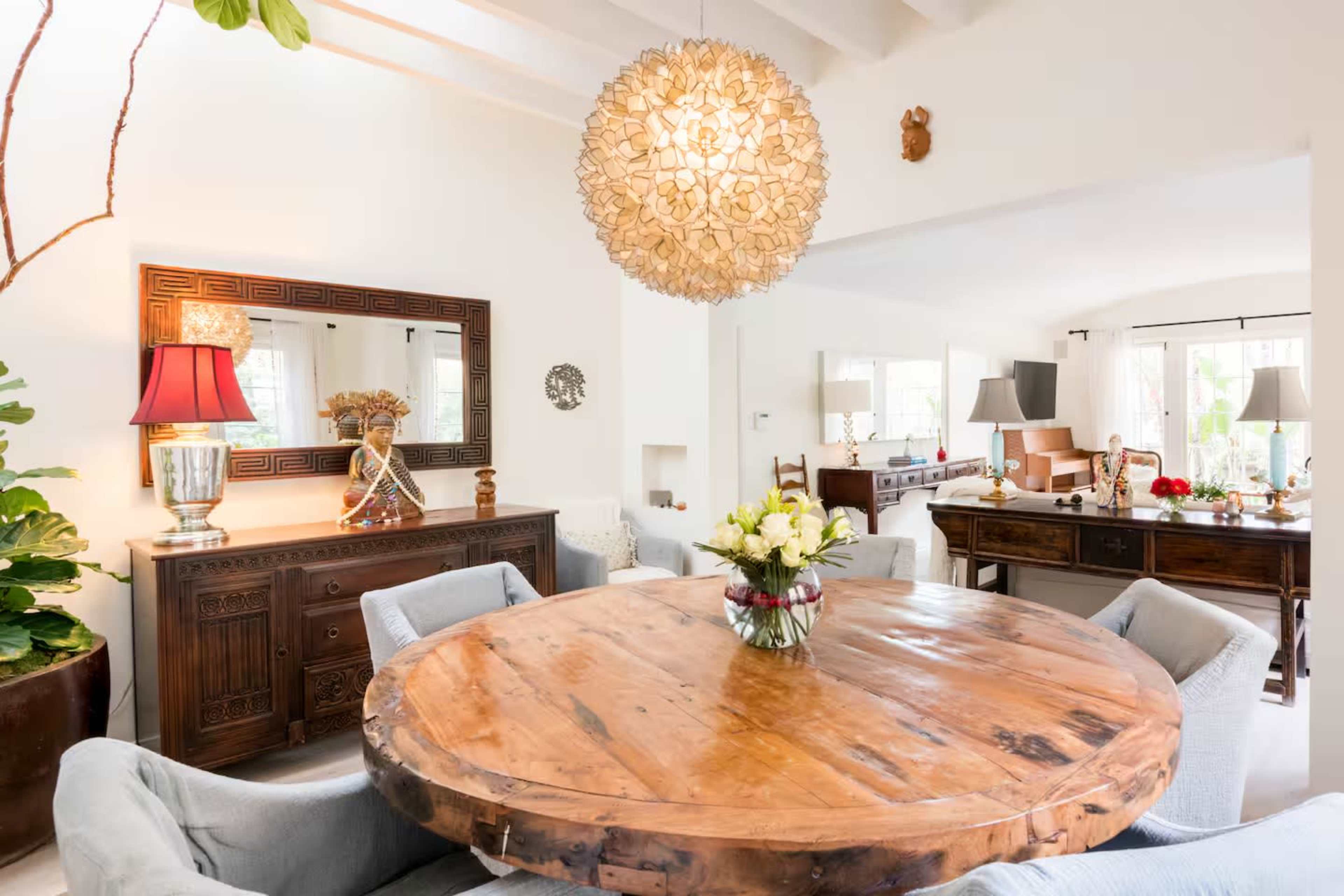 A round wooden dining table adorned with a floral centerpiece sits in a bright room filled with plants and decorative furniture.